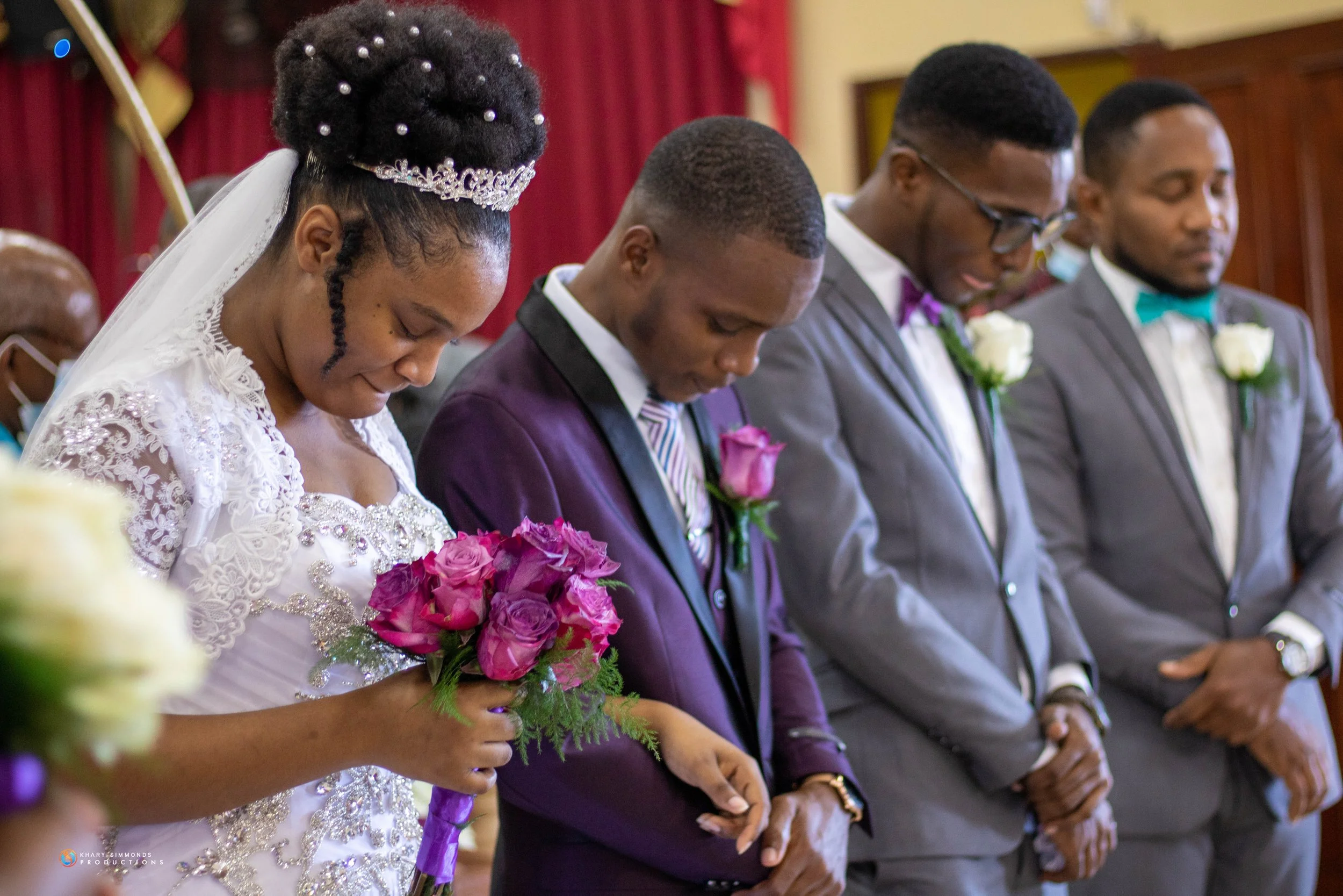 A bride in a white lace wedding dress with a tiara holding a bouquet of pink roses, and three groomsmen in suits with boutonnieres standing in prayer during a wedding ceremony.