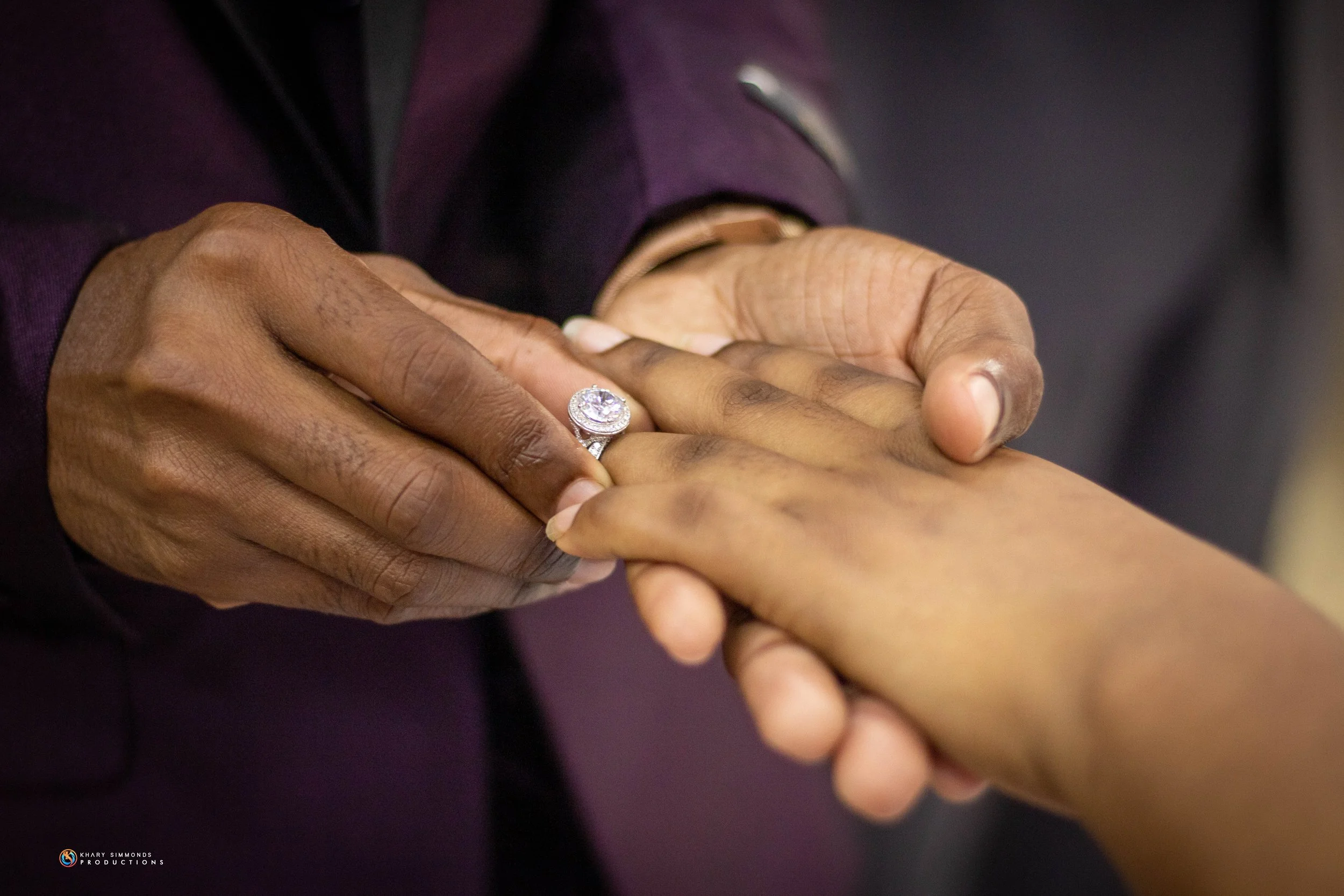Close-up of a man placing a diamond engagement ring on a woman's finger during a proposal, with both hands in focus against a blurred background.