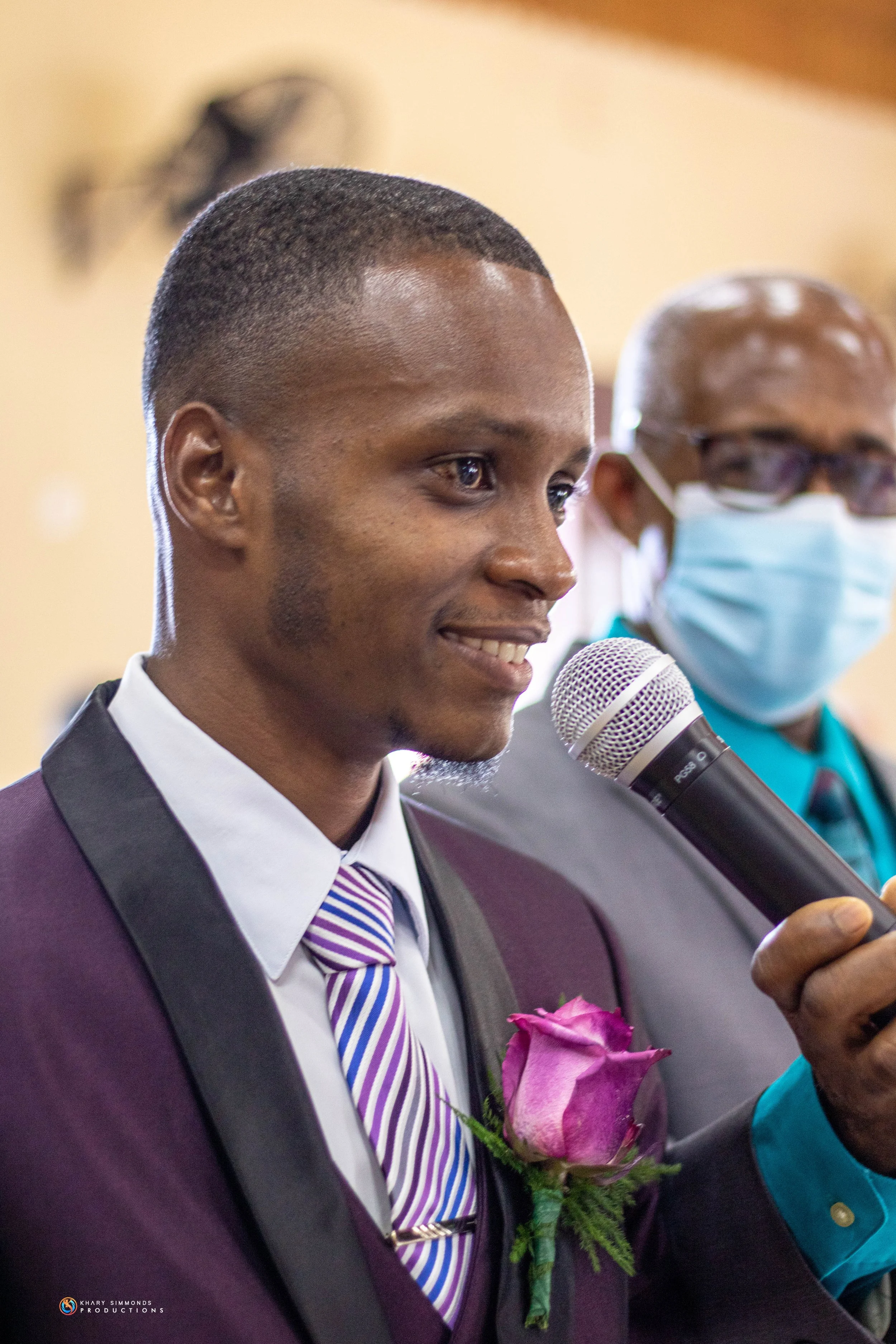 A young man in a suit with a purple boutonniere and a striped lavender tie is smiling and speaking into a microphone. Behind him, a man wearing glasses and a blue face mask is visible.