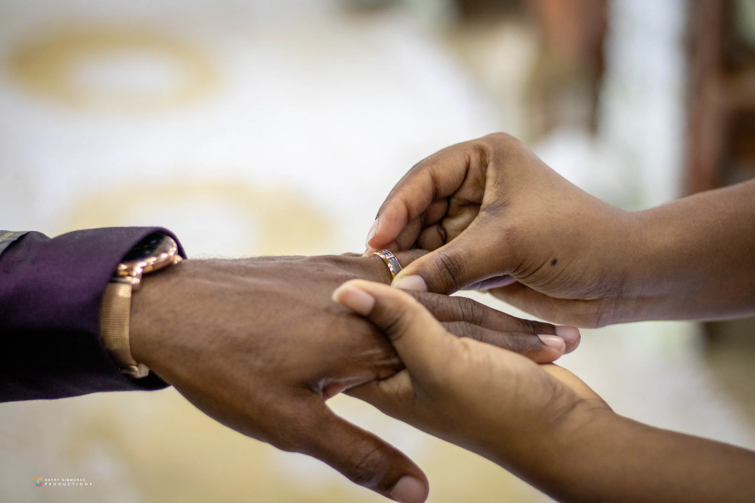A person places a wedding ring on another person's finger during a wedding or engagement ceremony, with a close-up focus on their hands.