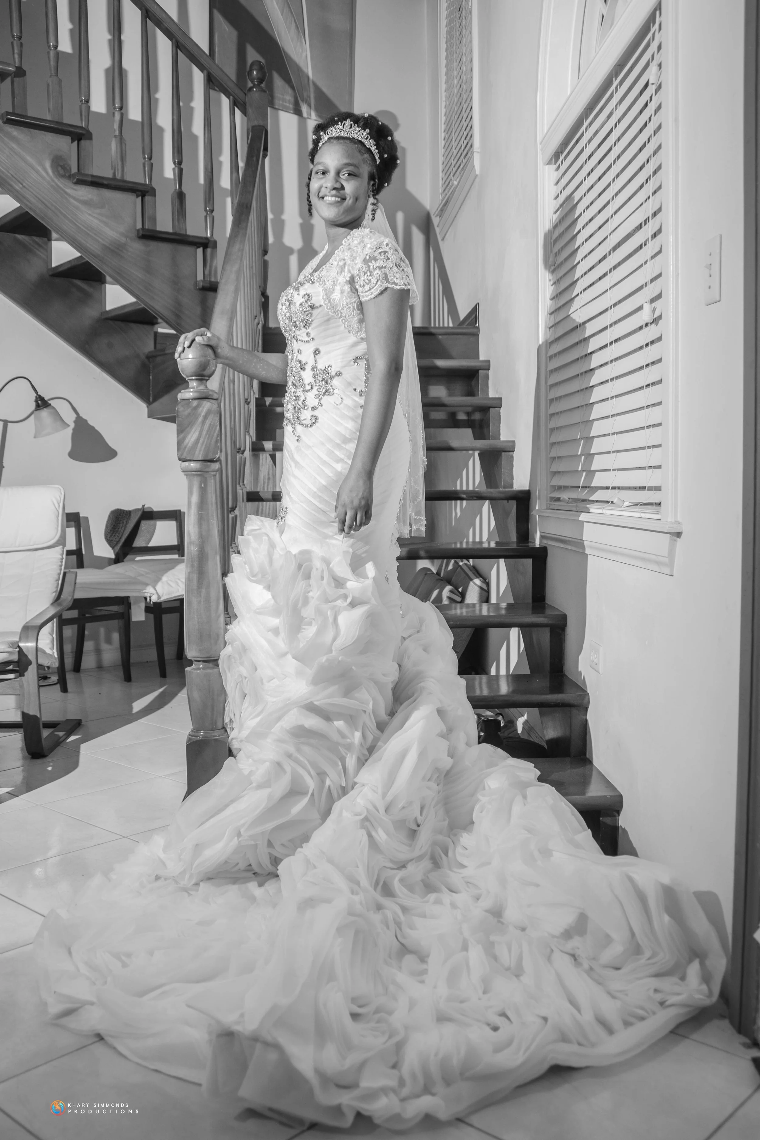 A woman in a wedding gown standing on a staircase, smiling, in a black-and-white photo.