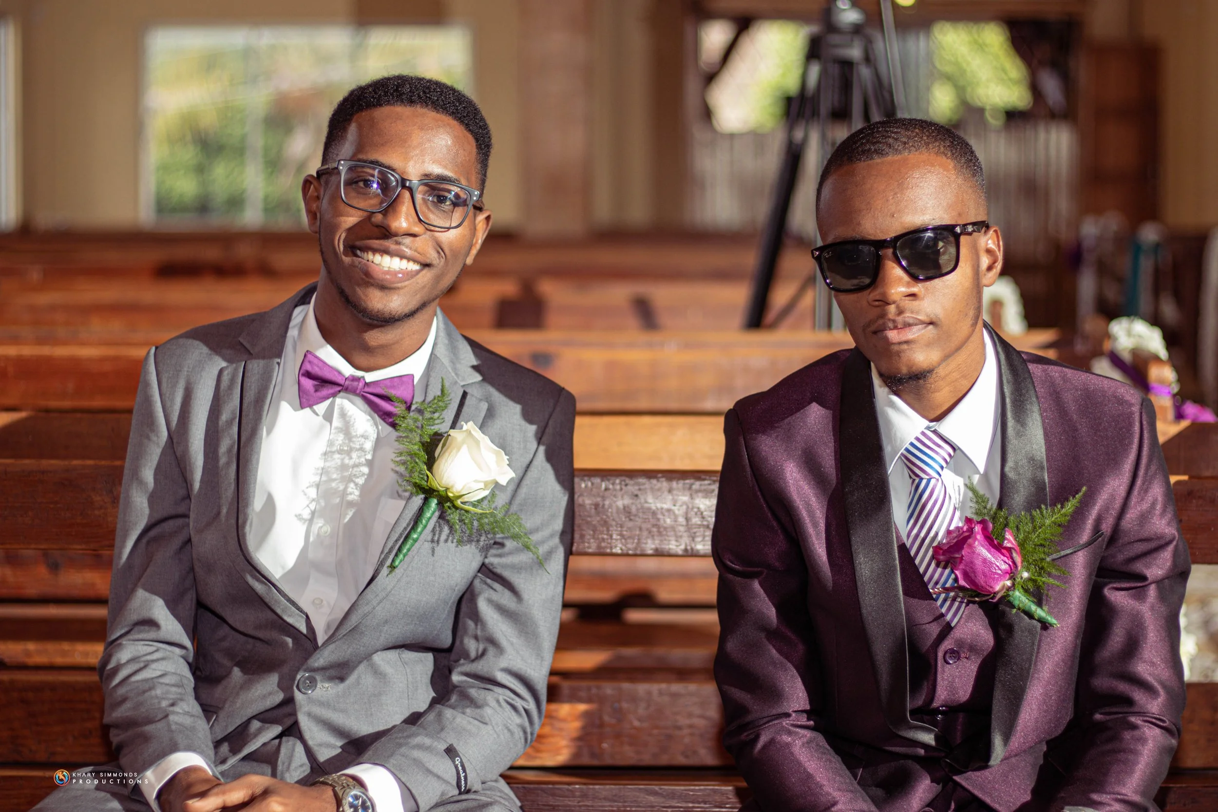 Two men in suits sitting on wooden benches inside a church, dressed for a wedding, both wearing glasses and boutonnières, with one smiling and the other looking serious.