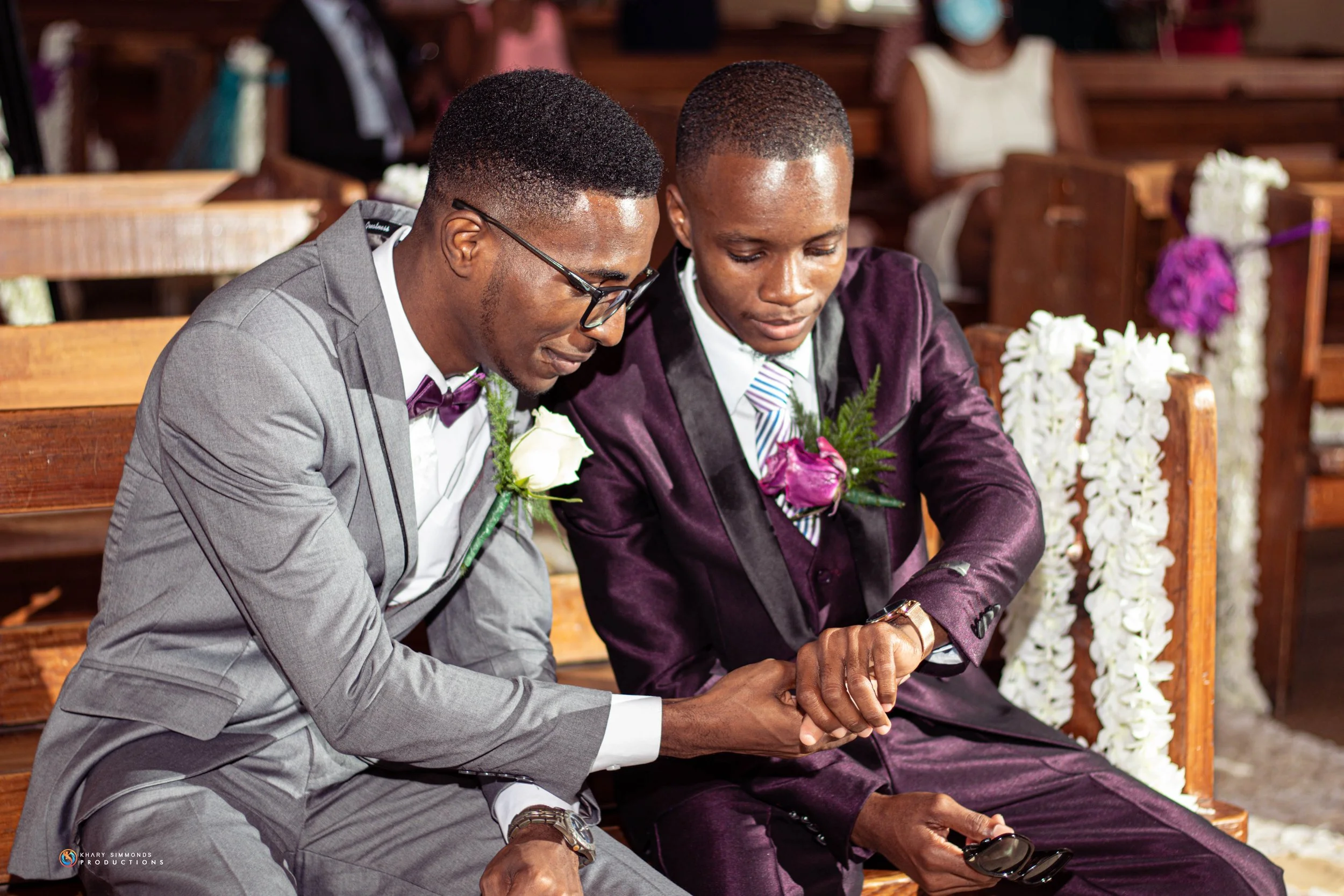 Two men in suits sitting on a wooden bench, holding hands and looking at the time on a watch. One is wearing a gray suit with a white shirt, purple bow tie, and glasses; the other is in a dark purple suit with a white and purple striped shirt. They a