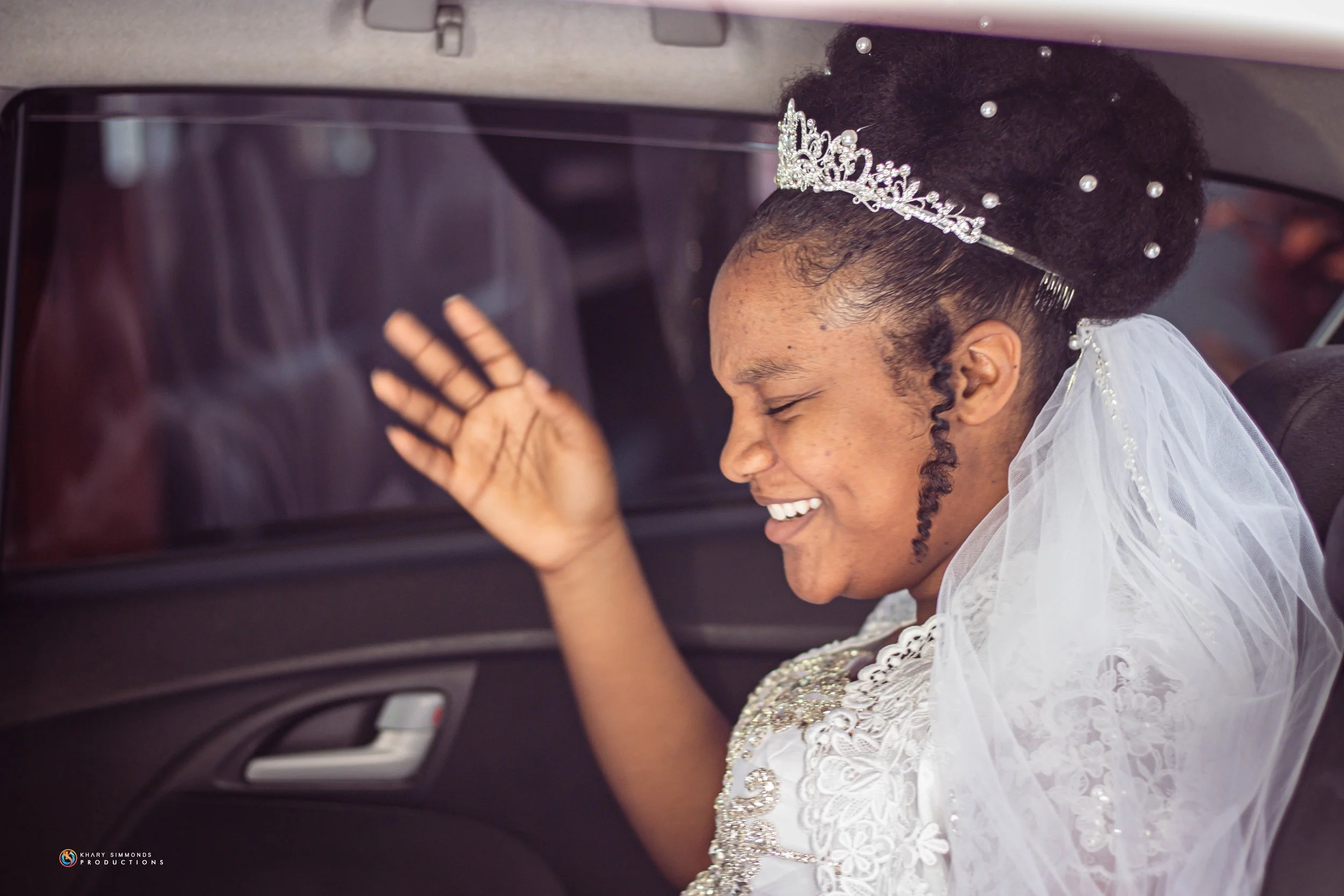 A bride with an elaborate updo styled with a tiara and pearls, smiling and waving while sitting inside a car, wearing a white lace wedding dress and veil.