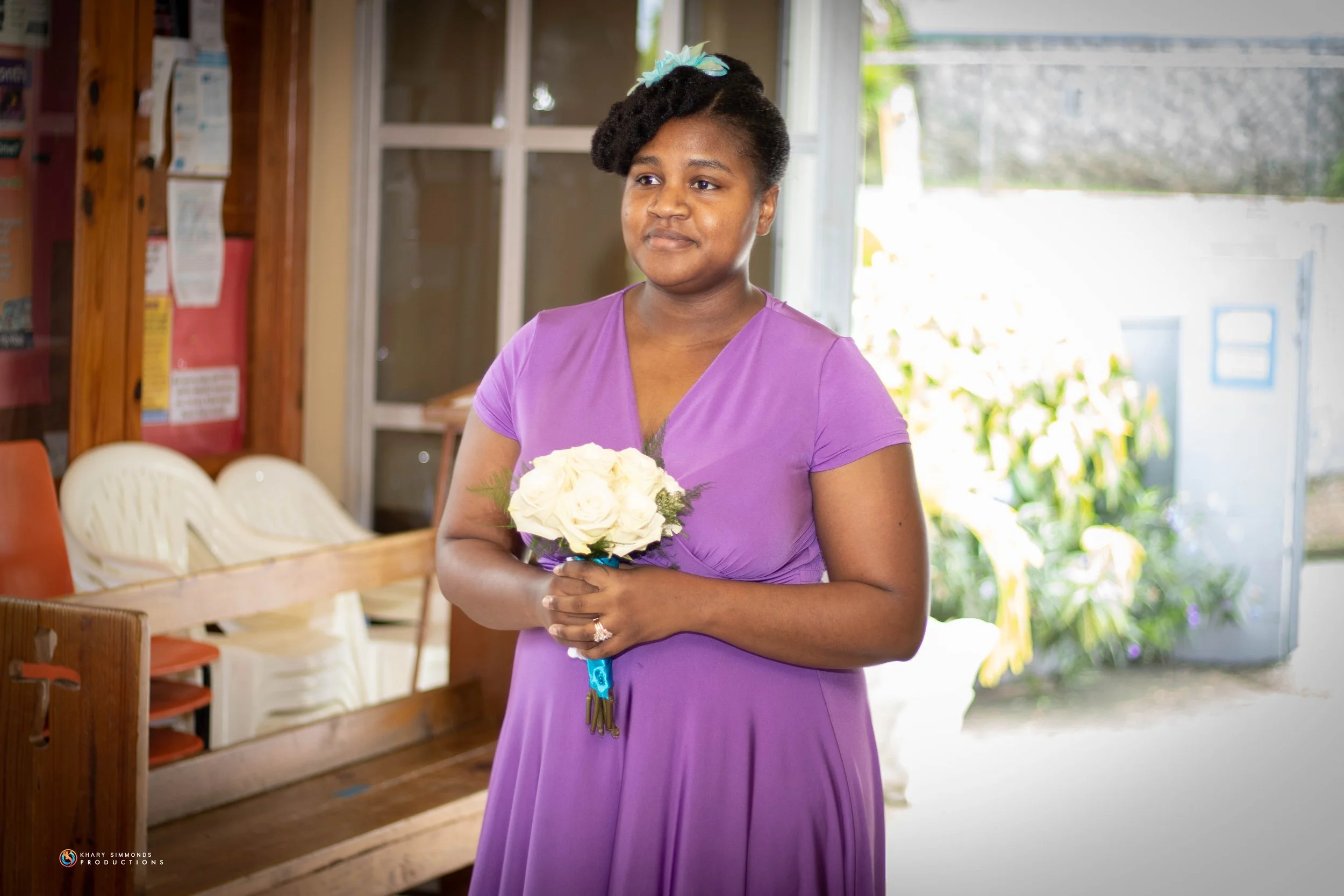 A woman in a purple dress holding a bouquet of white roses, standing indoors with a wooden bench and chairs behind her.