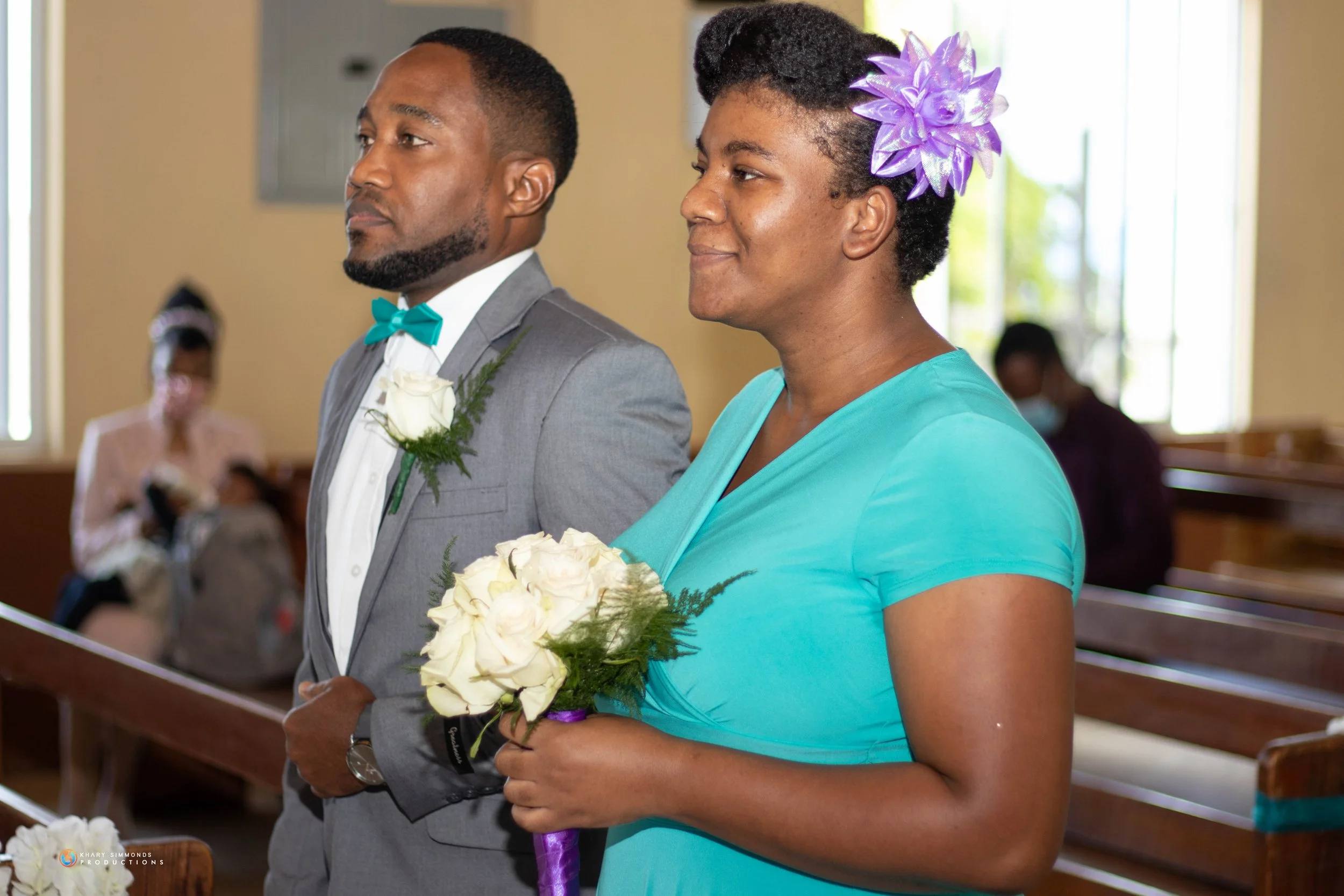 A man in a gray suit with a turquoise bow tie and white boutonniere standing next to a woman in a turquoise dress with a purple flower hair accessory, holding a bouquet of white roses, inside a church.