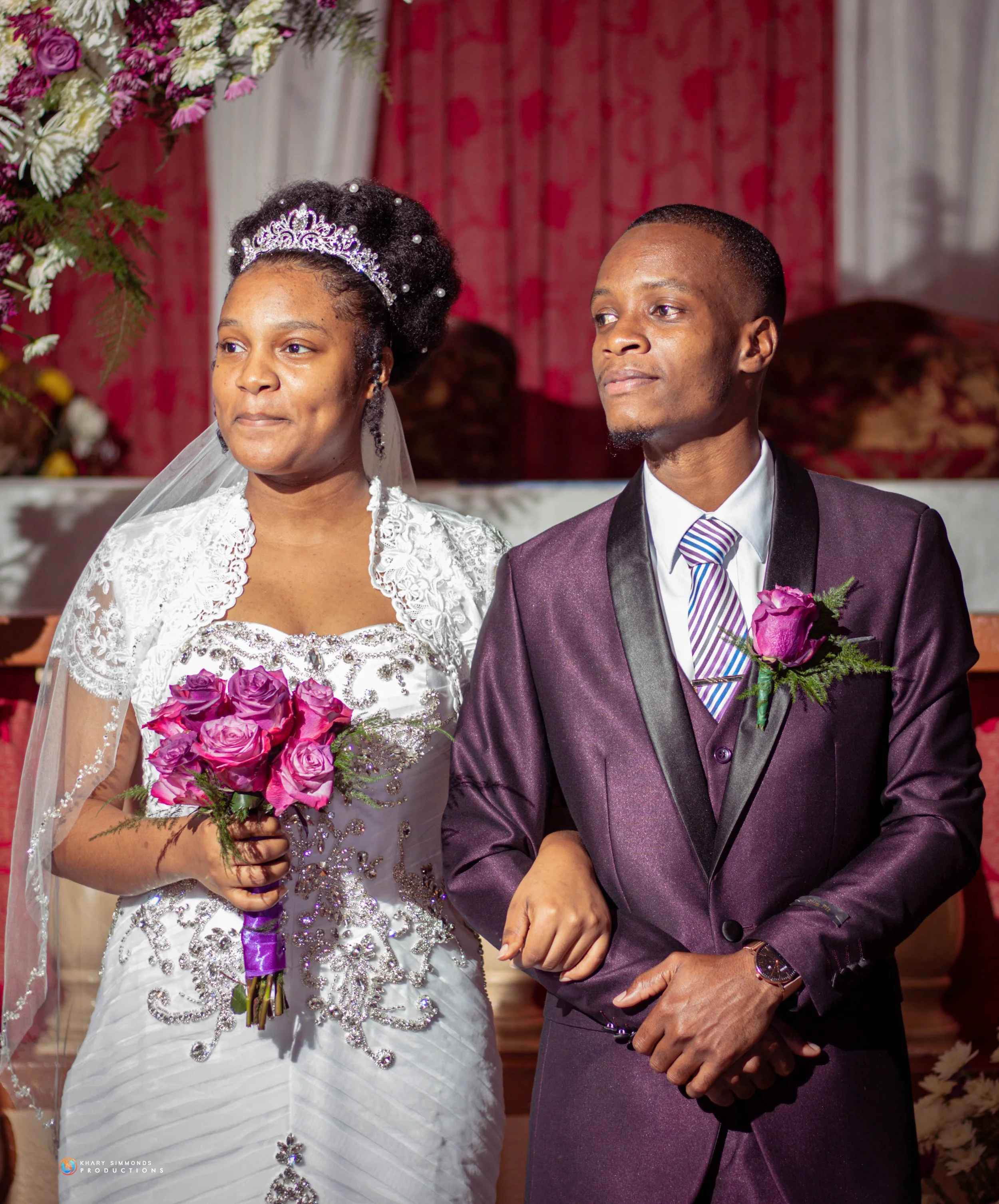 A bride and groom during their wedding ceremony, with both wearing formal attire and holding pink roses.