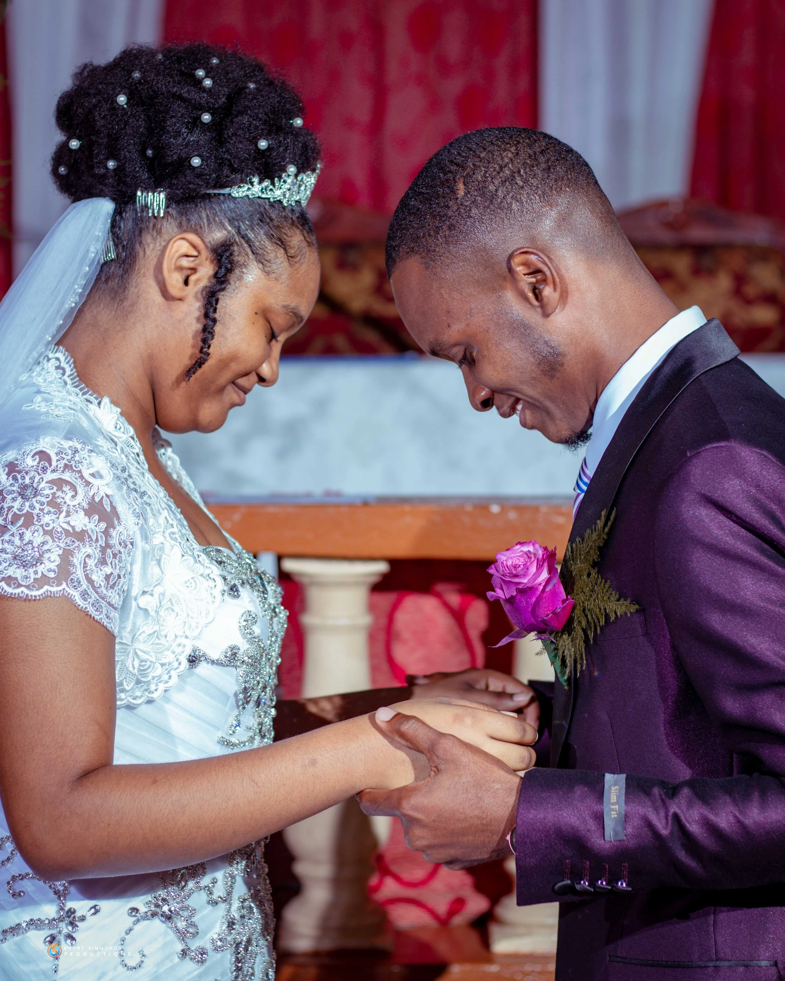 A bride and groom holding hands and bowing their heads during a wedding ceremony, with the bride wearing a white lace dress and the groom in a suit with a pink rose boutonniere.