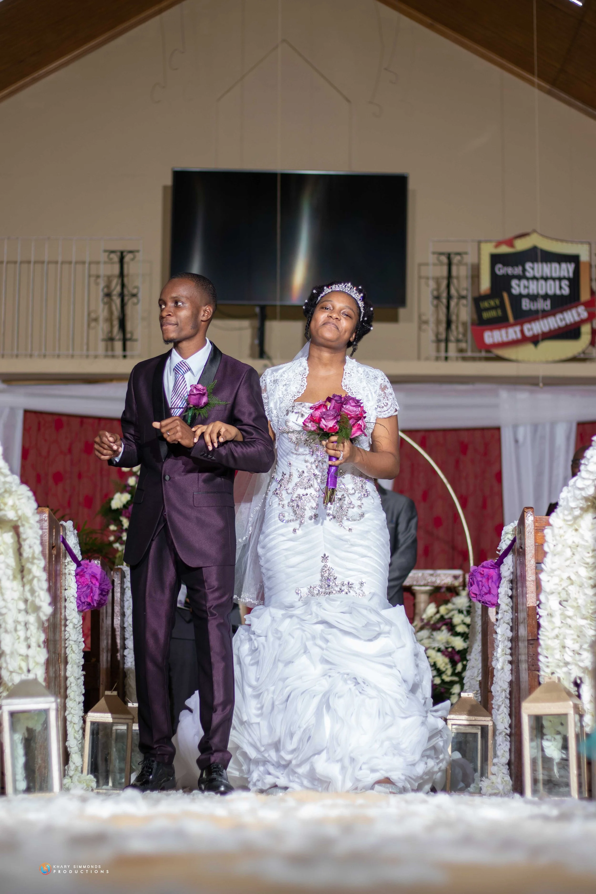 A bride and groom standing at the altar during a wedding ceremony inside a church, holding bouquets of pink flowers, with floral decorations around them.