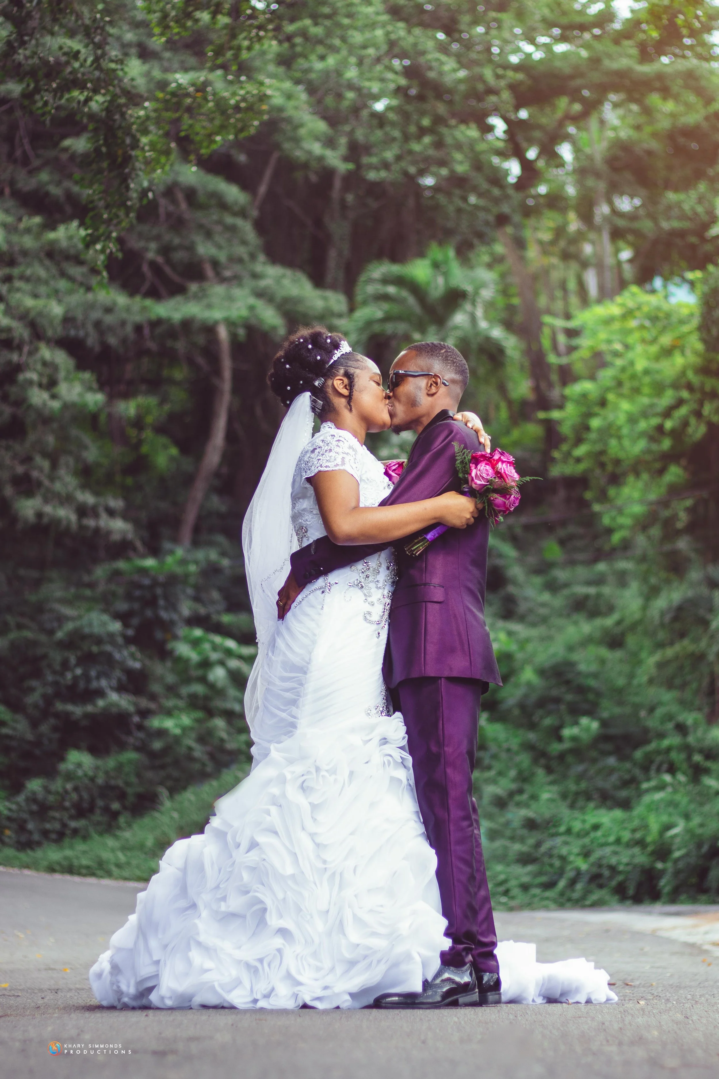 A newlywed couple sharing a kiss in a lush green outdoor setting, with the bride in a white wedding gown holding a bouquet of pink roses and the groom in a dark purple suit.