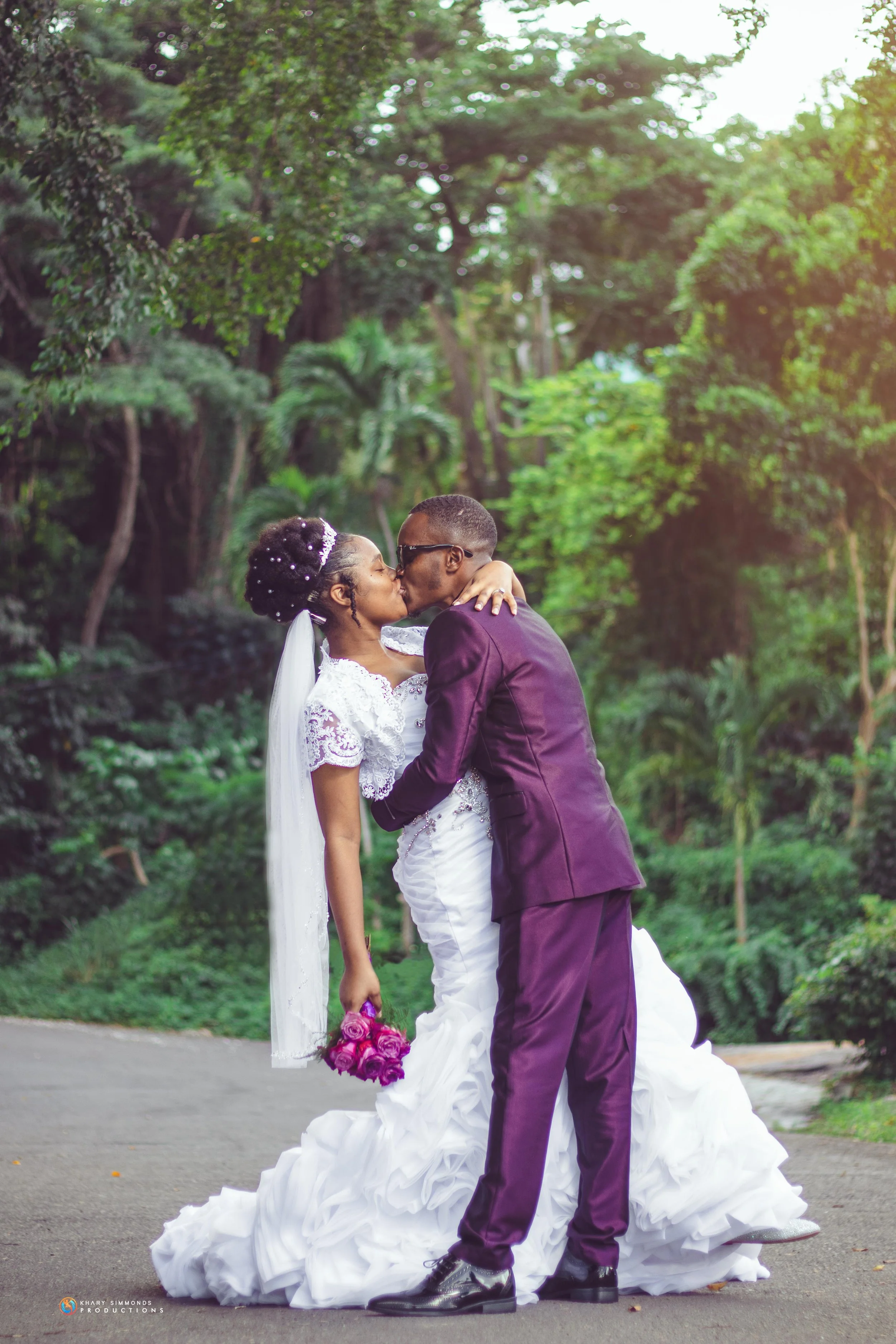 A bride and groom sharing a kiss outdoors on their wedding day, surrounded by green trees, with the bride holding a bouquet of pink roses.