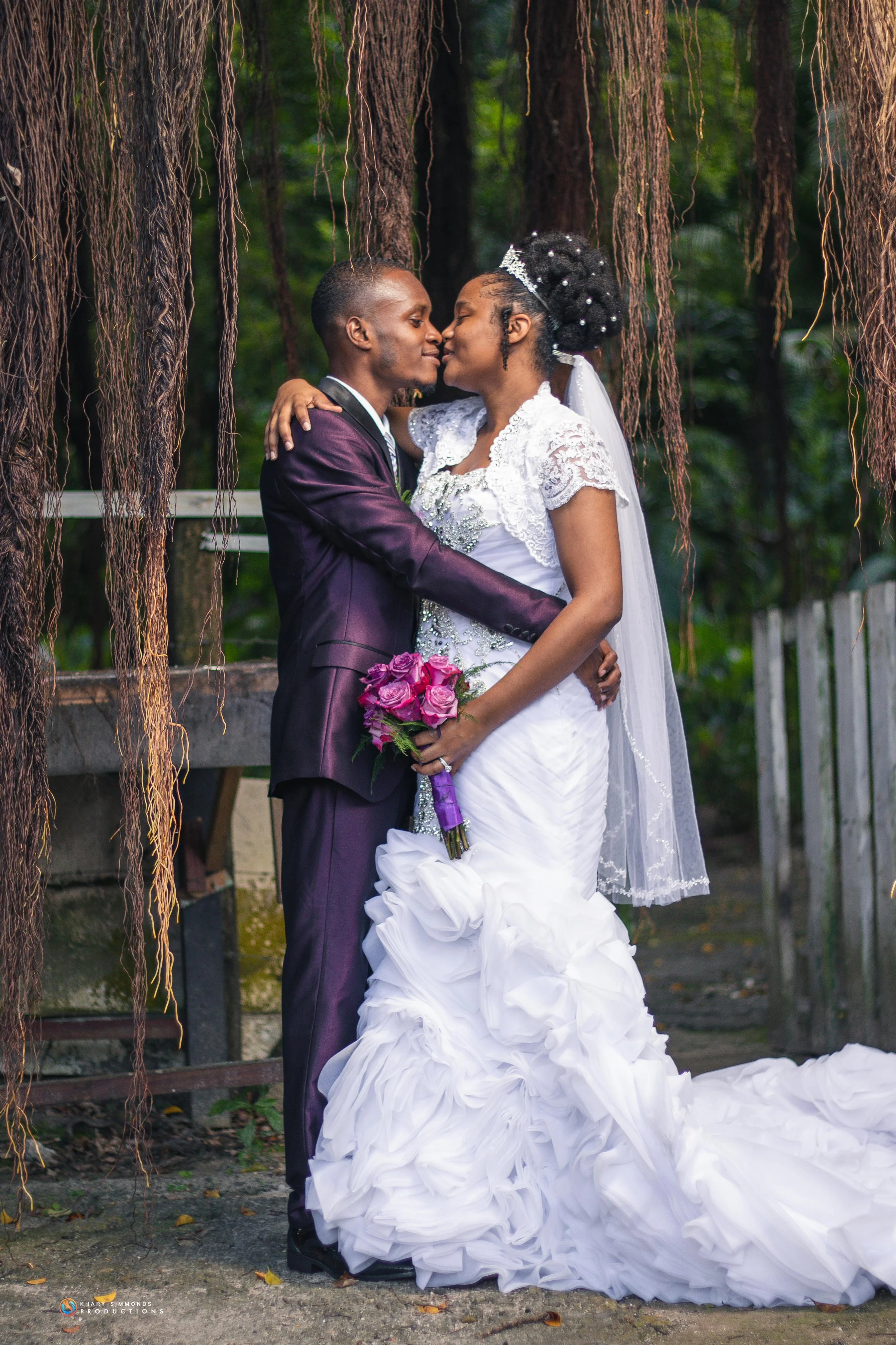 A bride and groom sharing a kiss outdoors, with the bride in a white wedding gown holding a bouquet of pink roses, and the groom in a dark suit, standing amidst hanging vines and greenery.