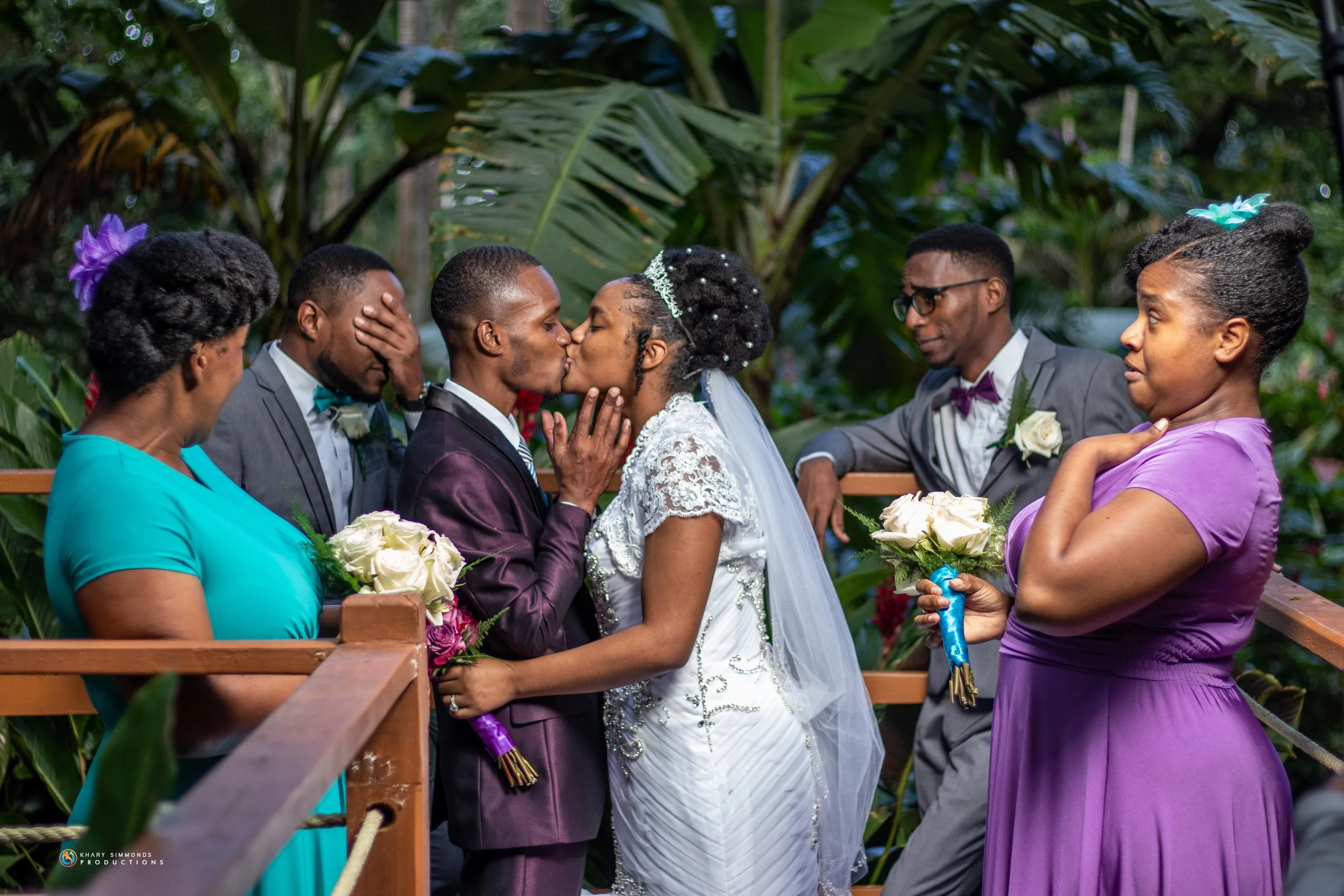 A wedding ceremony with the bride and groom sharing a kiss, surrounded by family members in formal attire, in a lush, tropical setting.