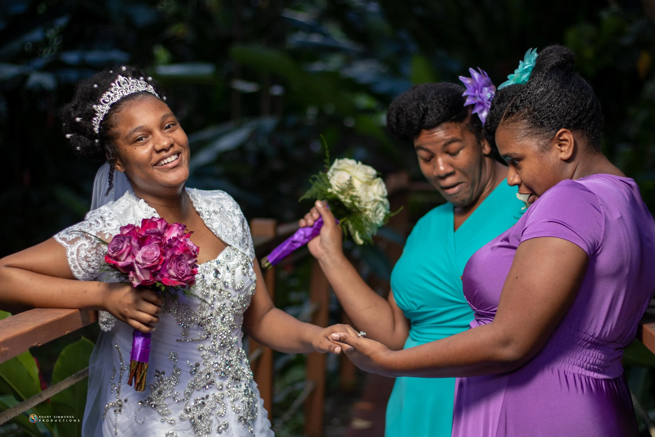 A bride in a white lace dress with a tiara and sash, holding a bouquet of pink roses, smiling as she holds hands with two women in a lush outdoor setting, one woman in a blue dress with a purple flower in her hair holding a white bouquet, and another