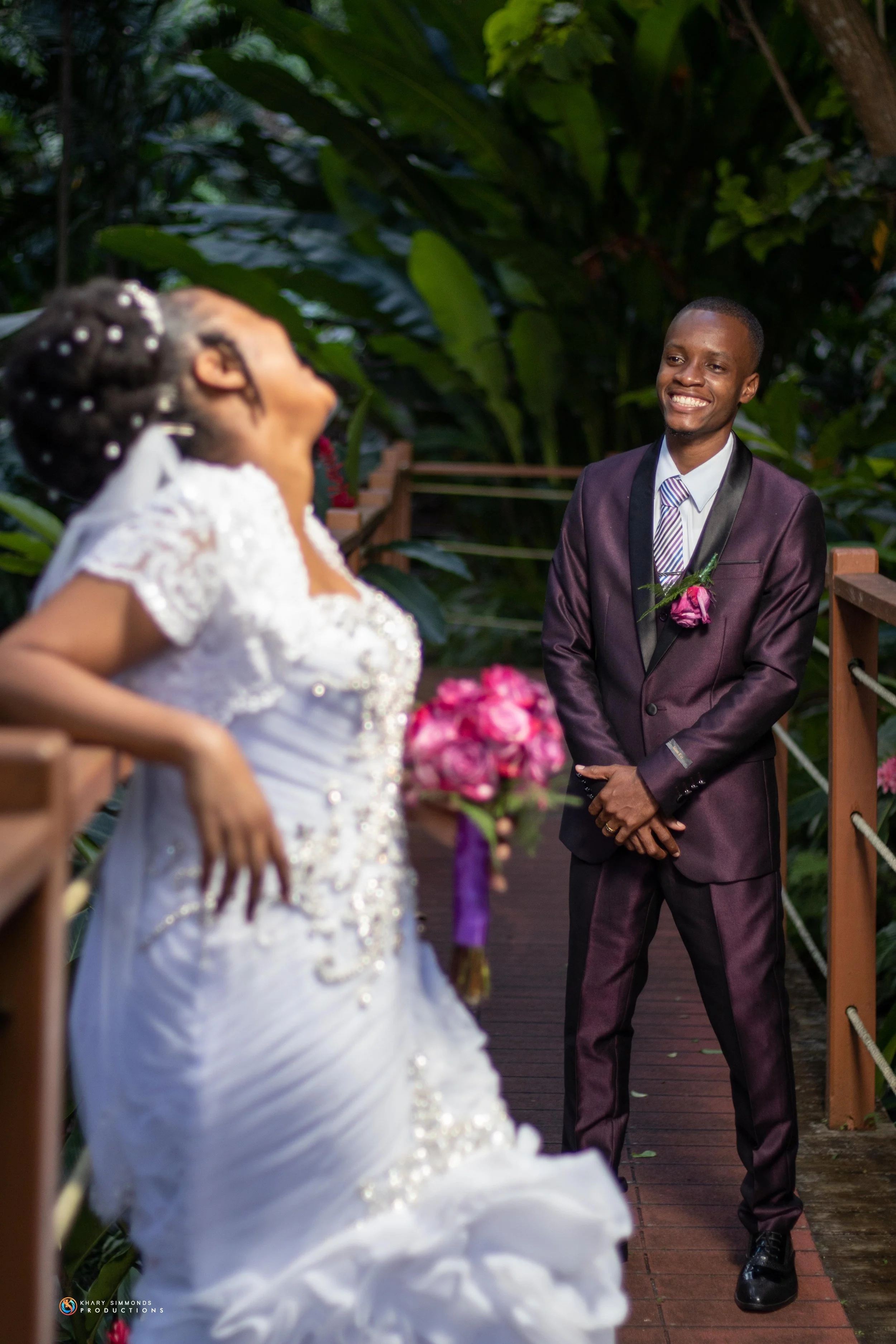 A newlywed couple in wedding attire sharing a joyful moment on a wooden bridge surrounded by lush green foliage. The bride is holding a pink bouquet and the groom is smiling at her.