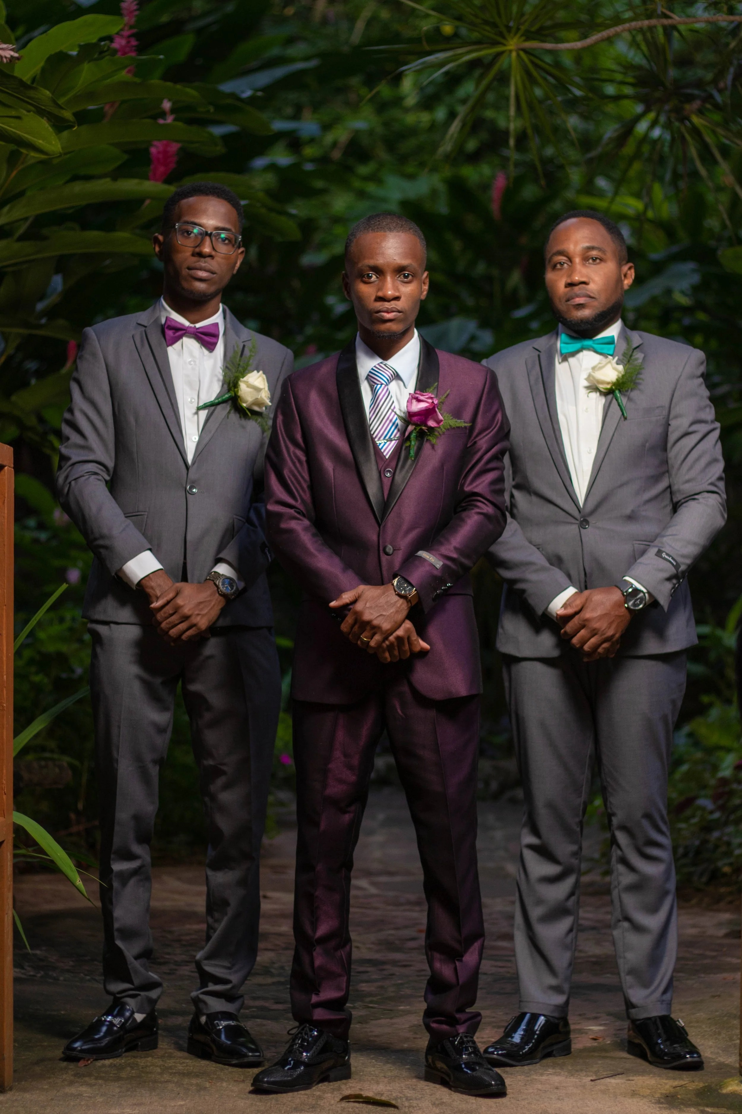 Three men in formal suits standing outdoors with greenery in the background.