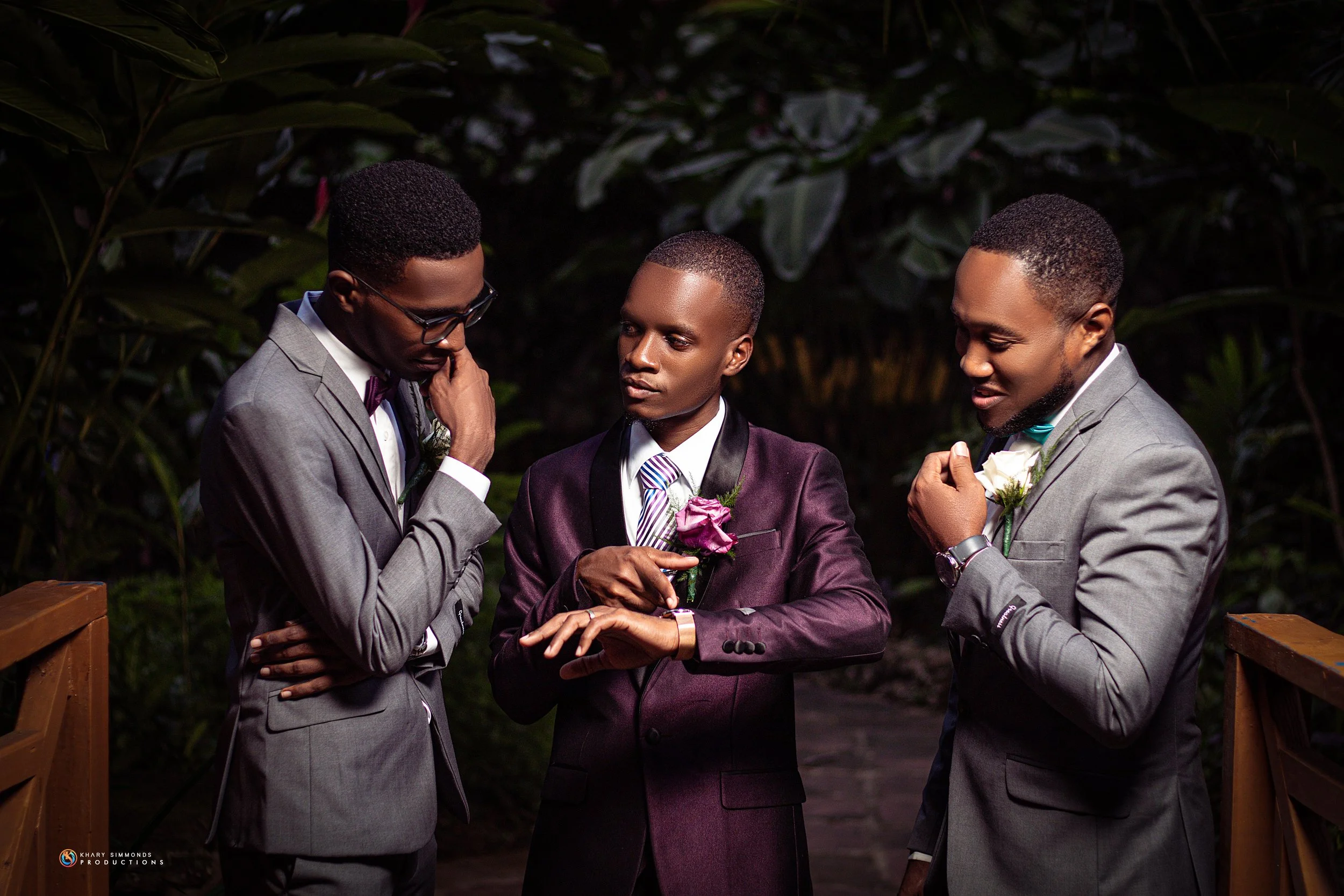 Three men in suits with boutonnières, standing outdoors at night, engaging in a conversation or ceremony, surrounded by foliage.