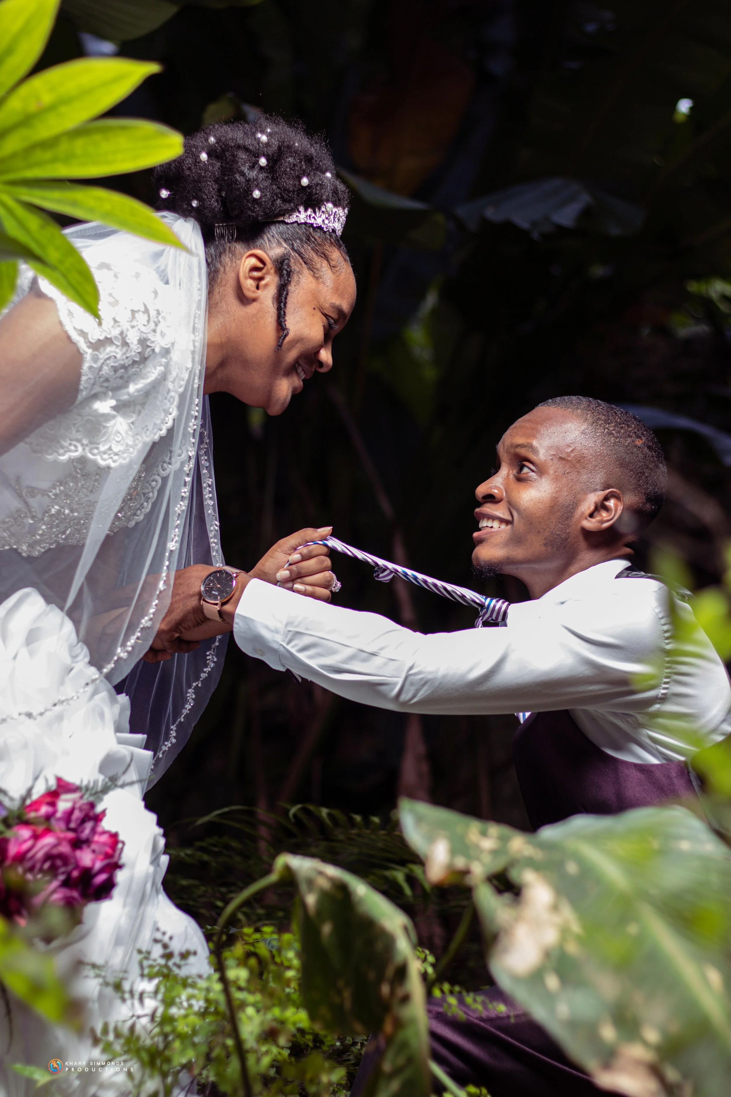 A couple on their wedding day, with the groom on one knee holding the bride's hands, smiling at each other outdoors surrounded by greenery.