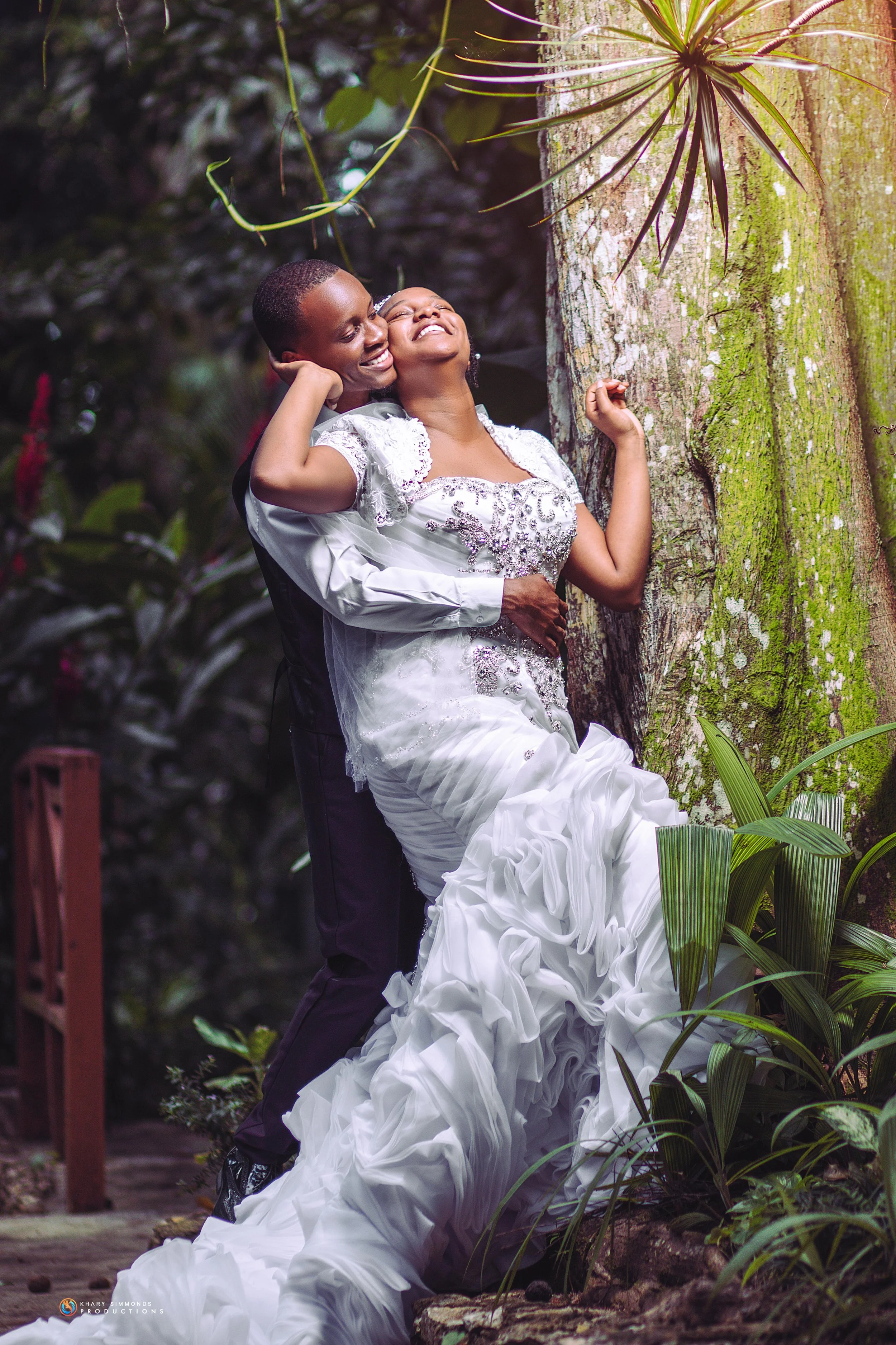 A joyful couple in wedding attire sharing a happy moment outdoors by a tree in a lush garden.