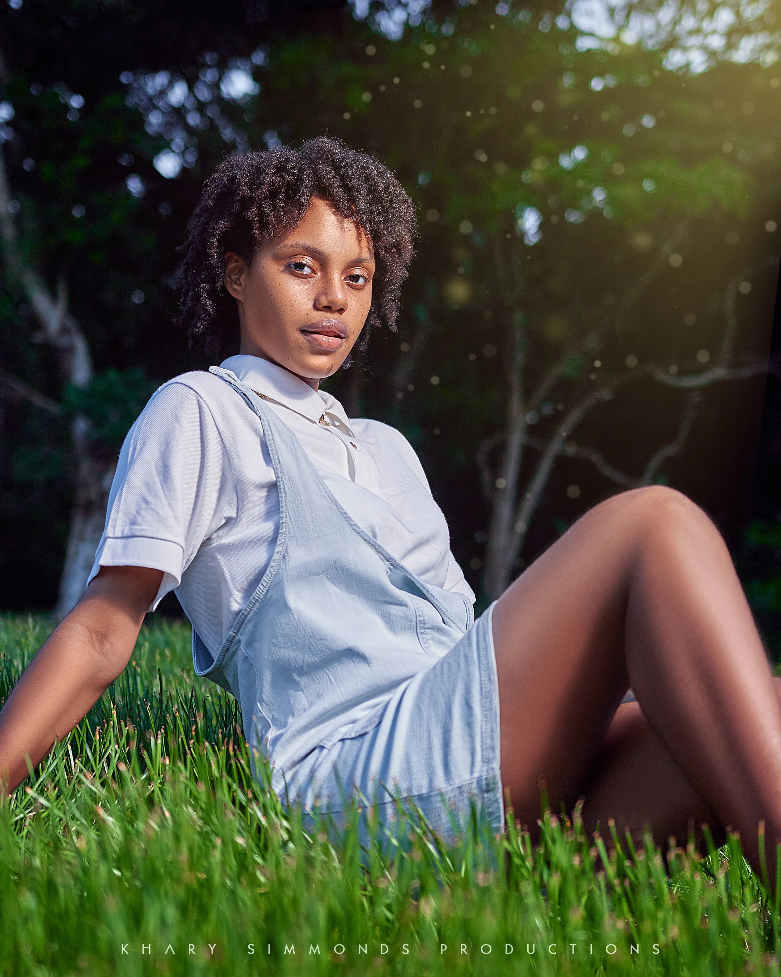 A young woman with natural curly hair sitting on grass outdoors, wearing a light-colored shirt and overalls, with trees and sunlight in the background.