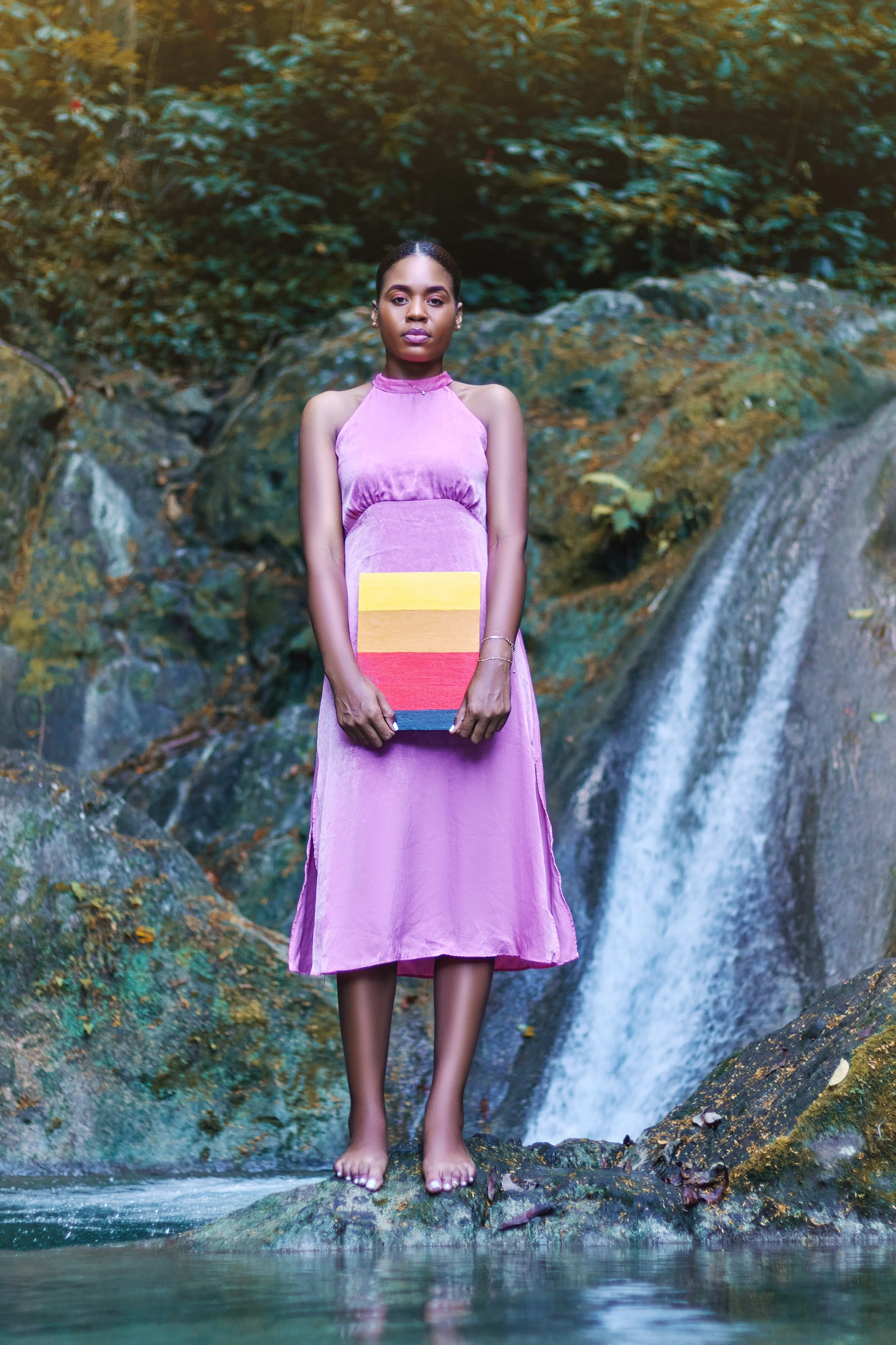A woman in a pink dress stands barefoot on rocks in front of a waterfall and lush greenery, holding a colorful striped clutch.