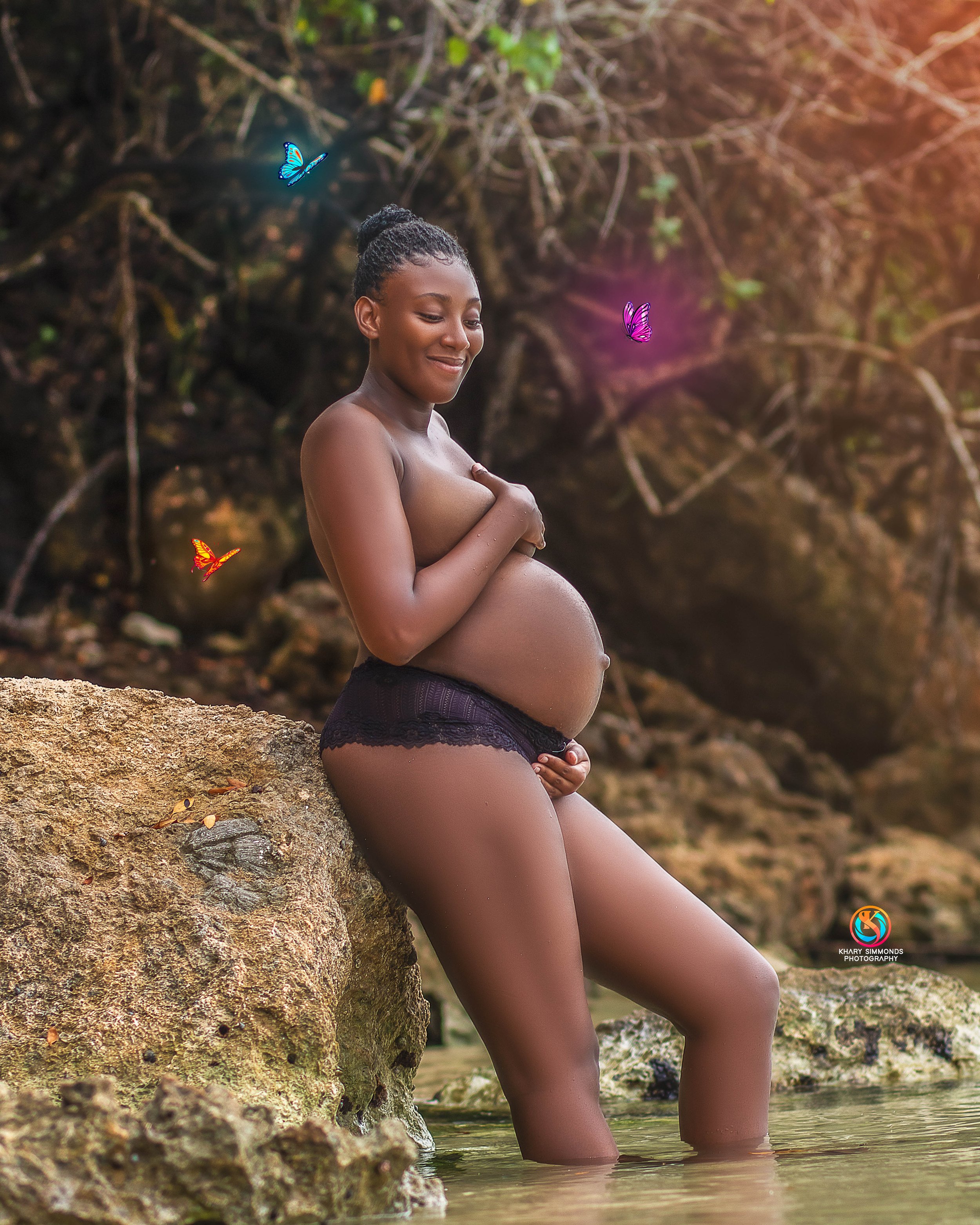 A pregnant woman with dark skin and curly hair tied up in a bun, sitting on a rock in a river. She is topless, wearing black lace underwear, with her knees in the water. She is smiling and touching her belly, surrounded by colorful butterflies in an 