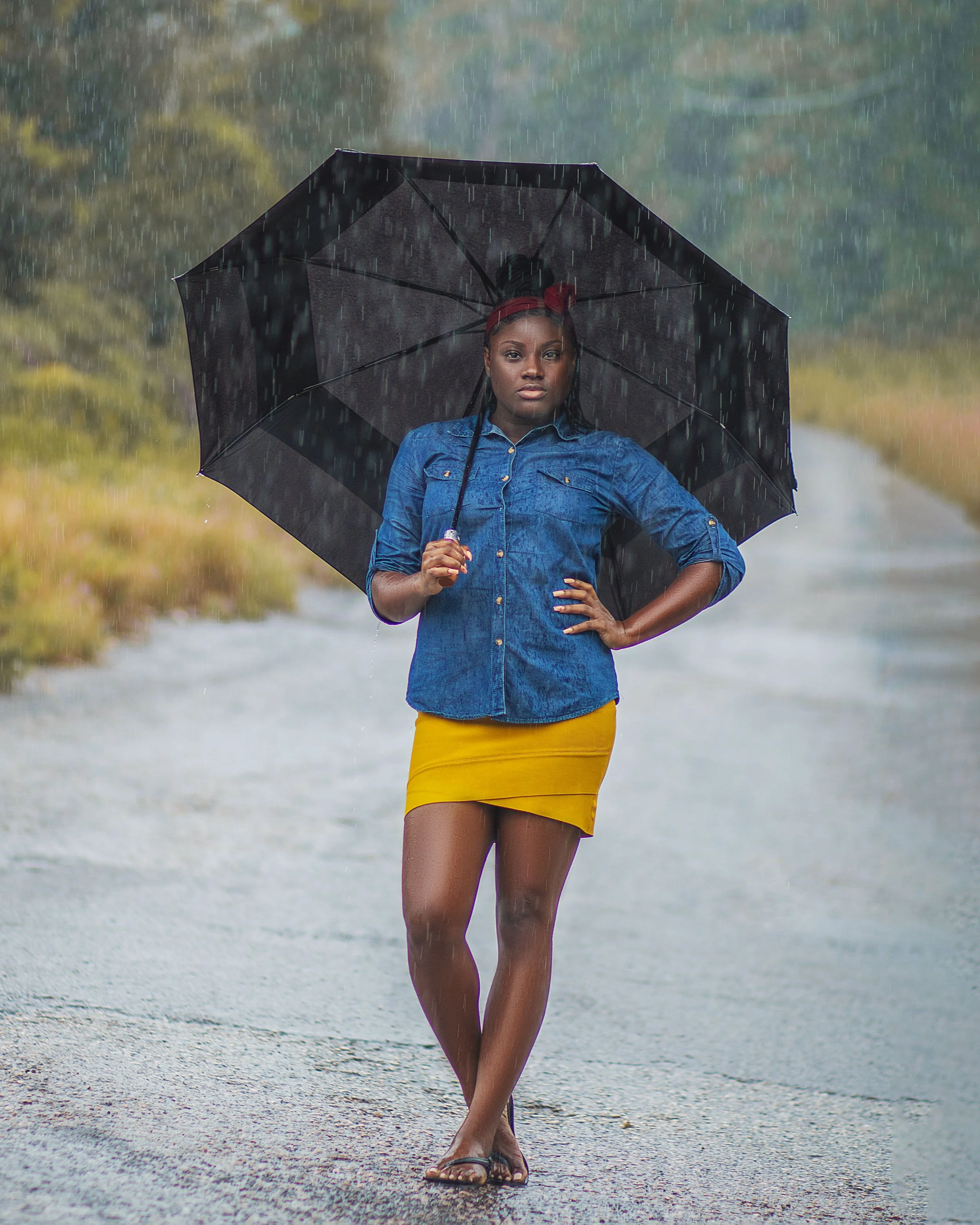 A woman standing on a rainy road, holding a black umbrella. She is wearing a blue denim shirt and a yellow skirt, with her hand on her hip and looking at the camera.