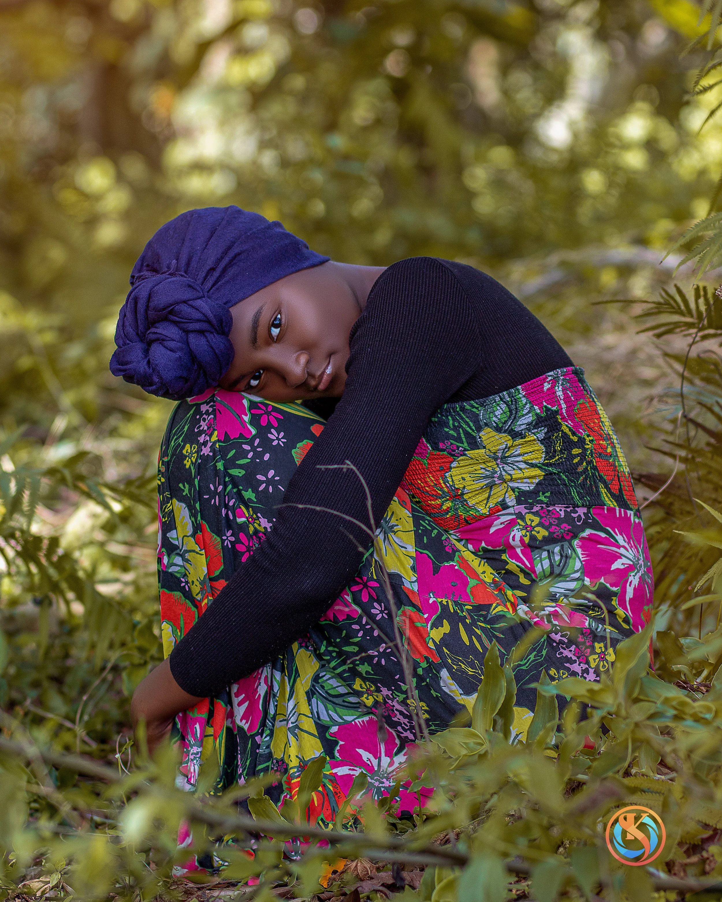 A woman with dark skin, wearing a purple headwrap, black top, and a vibrant floral skirt, sitting outdoors among green foliage with a contemplative expression.