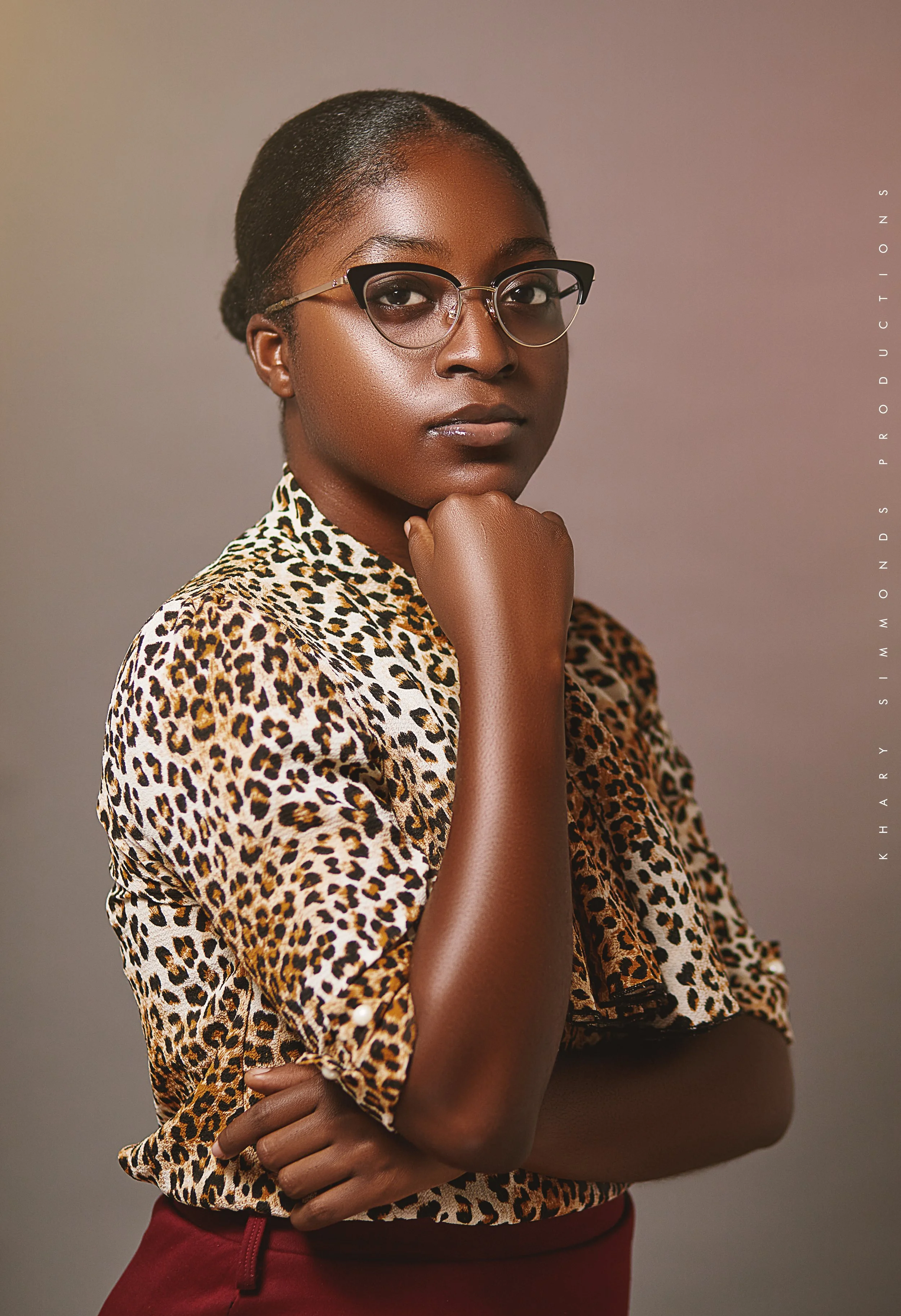 A woman wearing glasses and a leopard print blouse, posing against a plain background, with one hand resting under her chin.
