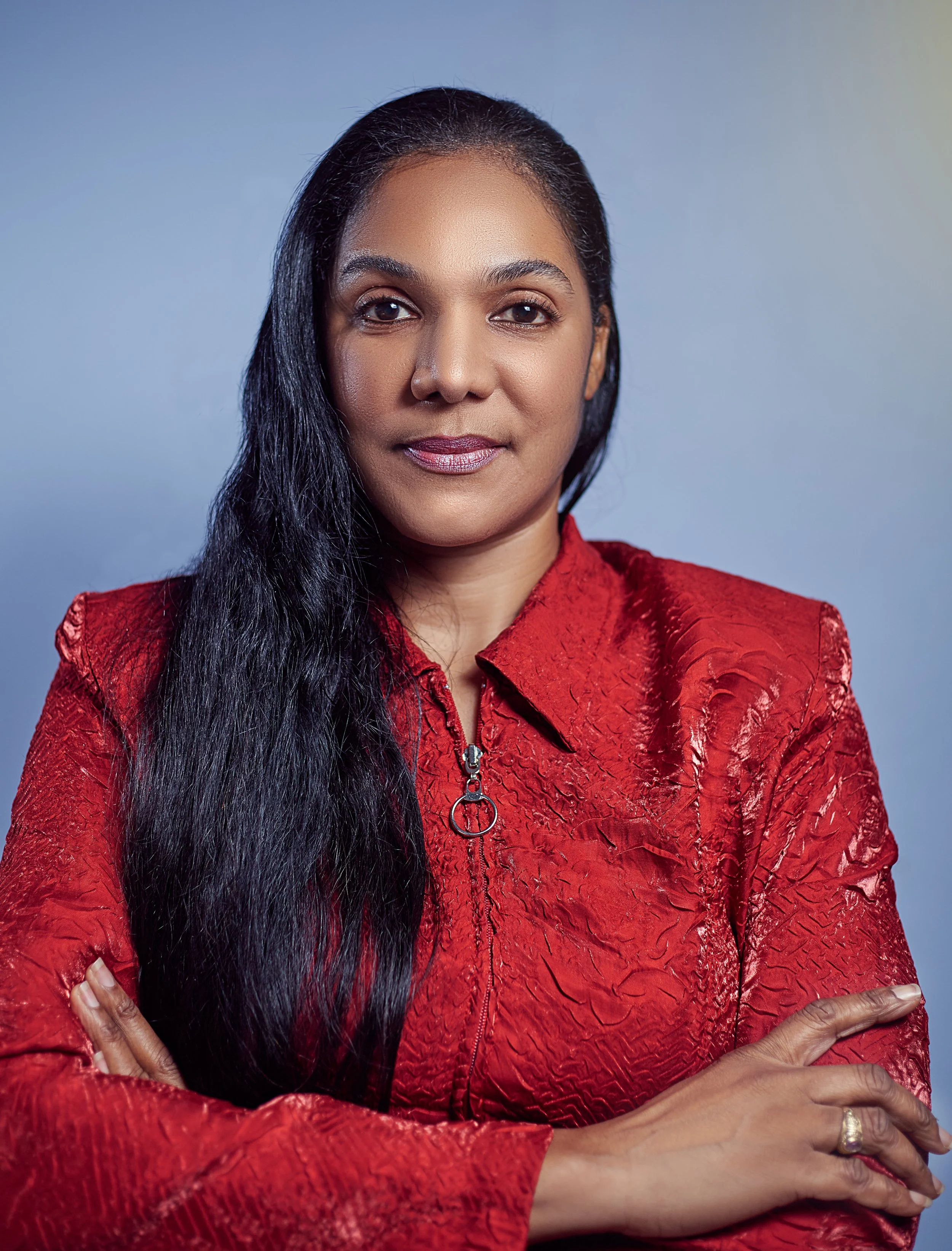 Portrait of a woman with long black hair, wearing a red textured jacket, arms crossed, against a blue background.