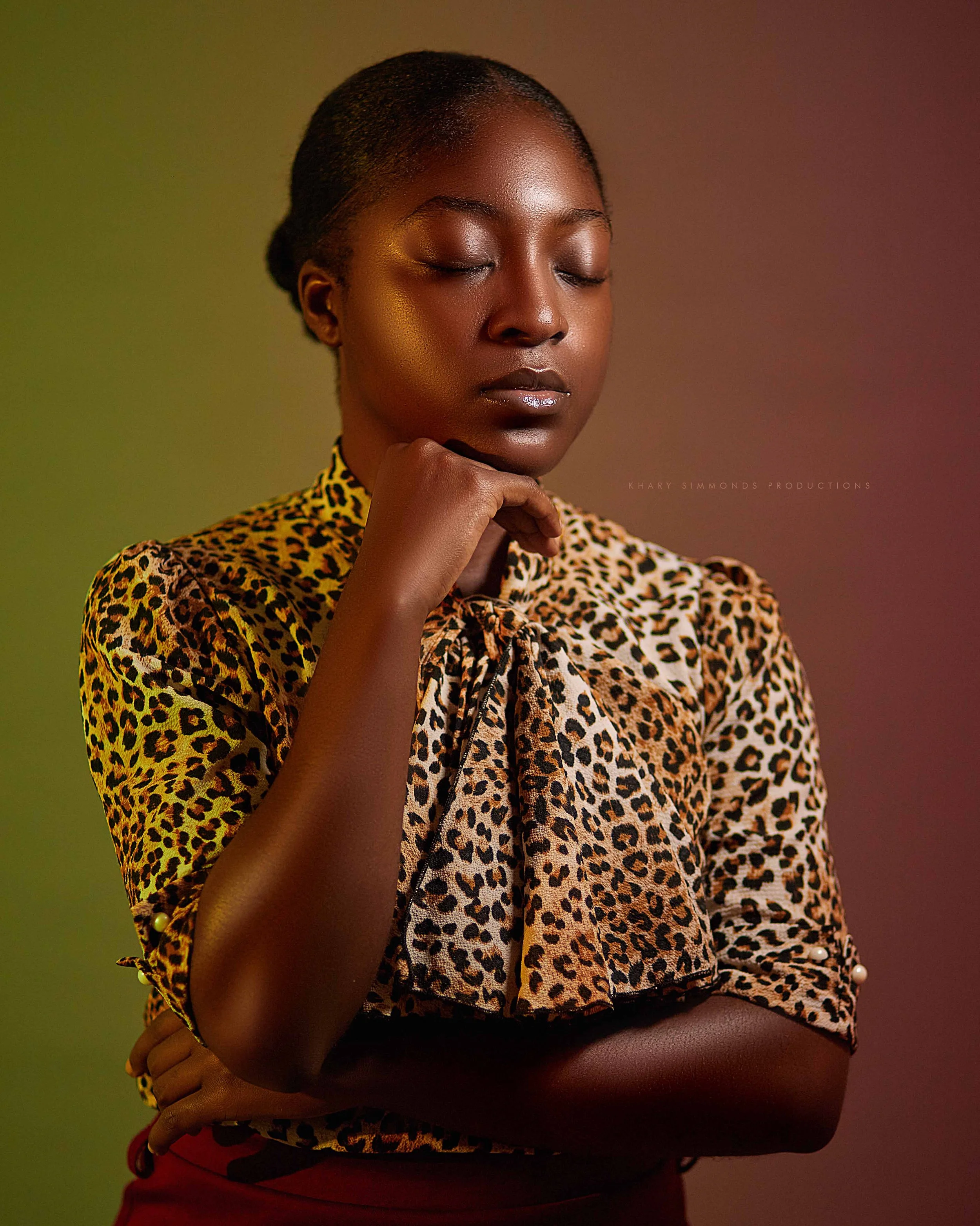 A woman with dark skin and her eyes closed, wearing a leopard print blouse, standing with her right hand resting on her chin and her left arm crossed over her waist.