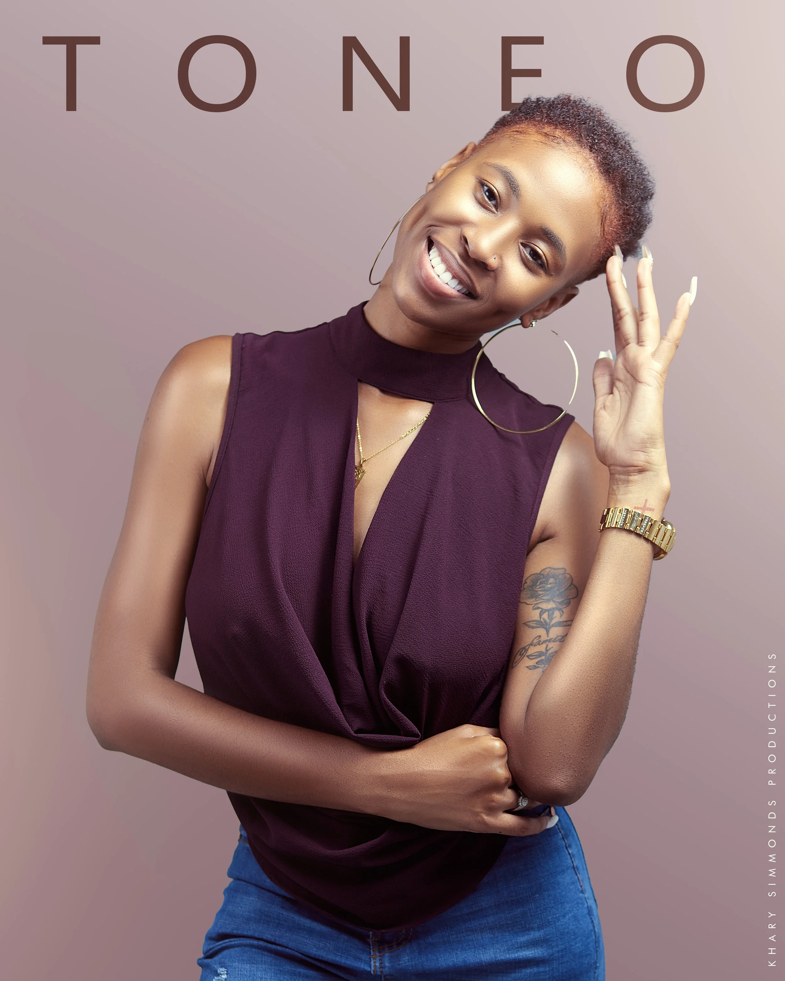 Smiling woman with short curly hair, wearing a sleeveless dark purple top, large hoop earrings, a gold watch, and a tattoo on her left arm, posing with one hand touching her head and the other arm crossed.