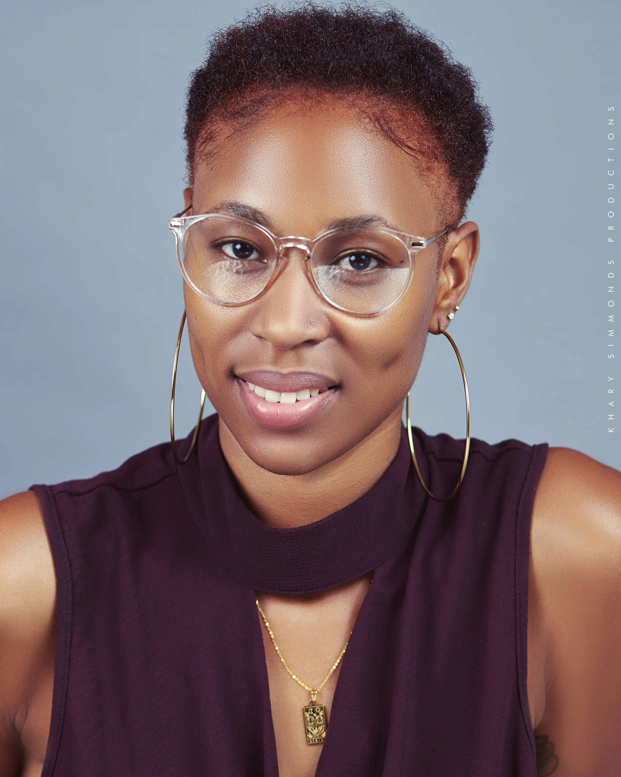 A young woman with short curly hair, wearing clear glasses, large hoop earrings, a purple sleeveless top, and a gold necklace with a pendant, smiling against a plain light blue background.