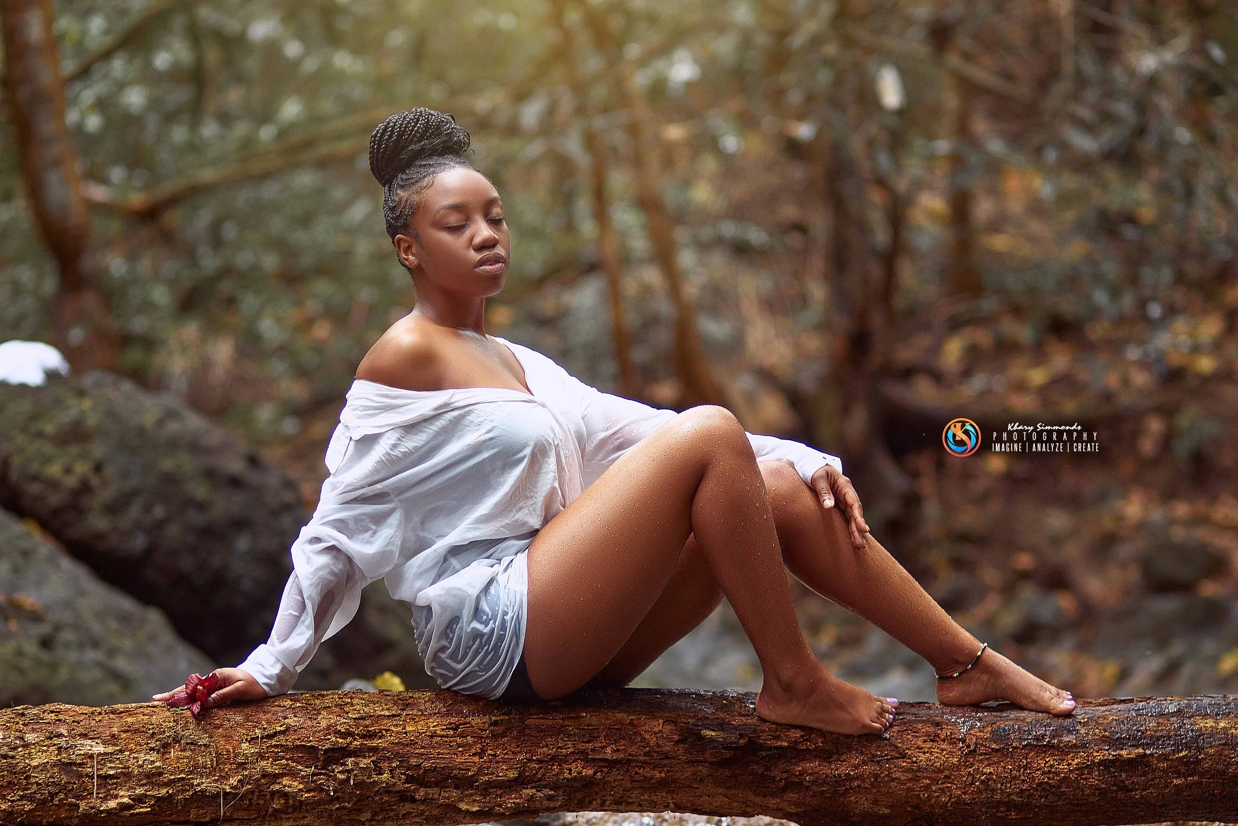 A woman with braided hair sitting on a fallen log in a forest, wearing a white off-shoulder shirt and shorts, with her eyes closed.