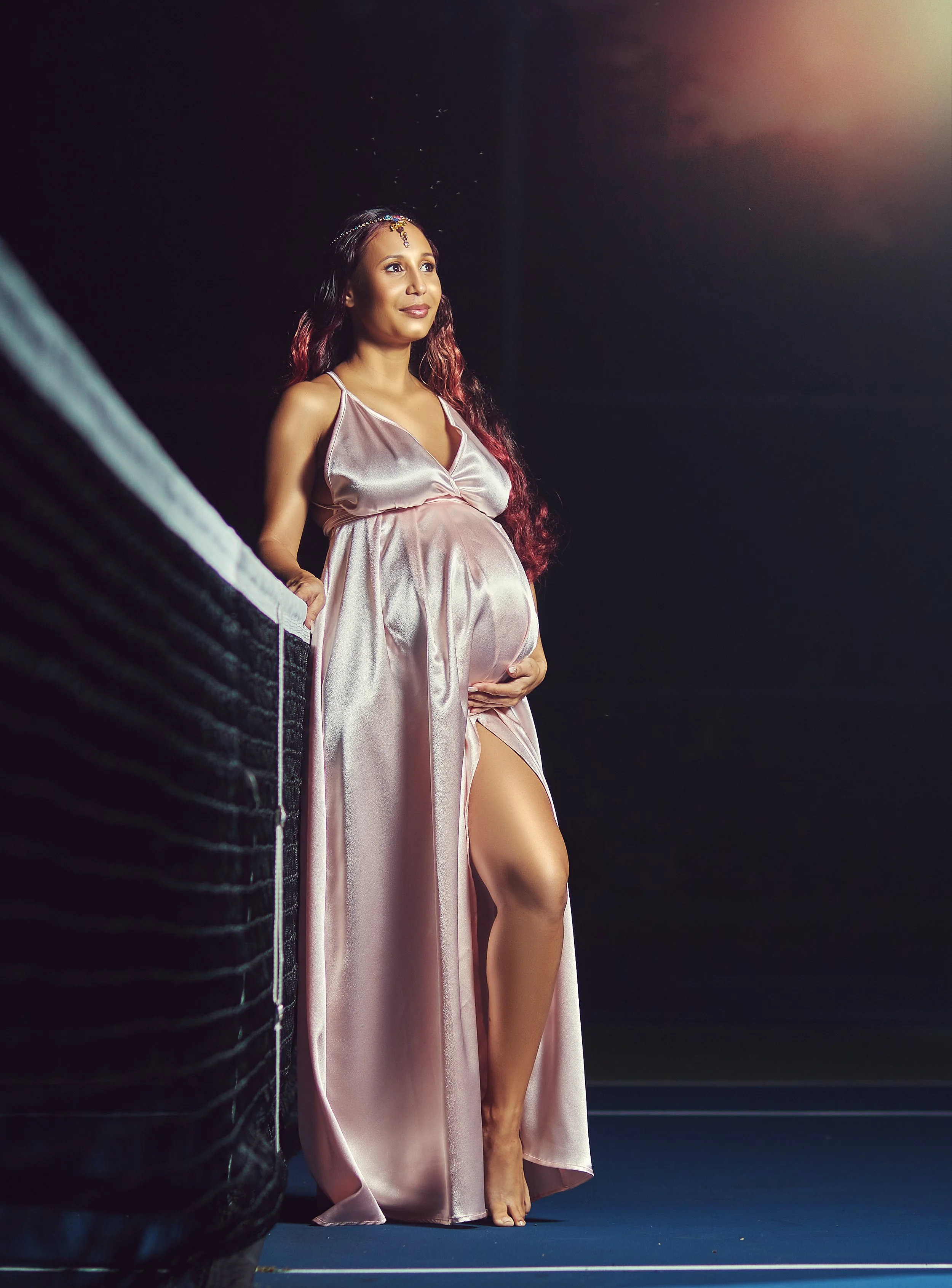 Pregnant woman in a silky pink gown standing barefoot on a dark stage, illuminated by a spotlight, with a black curtain as background.