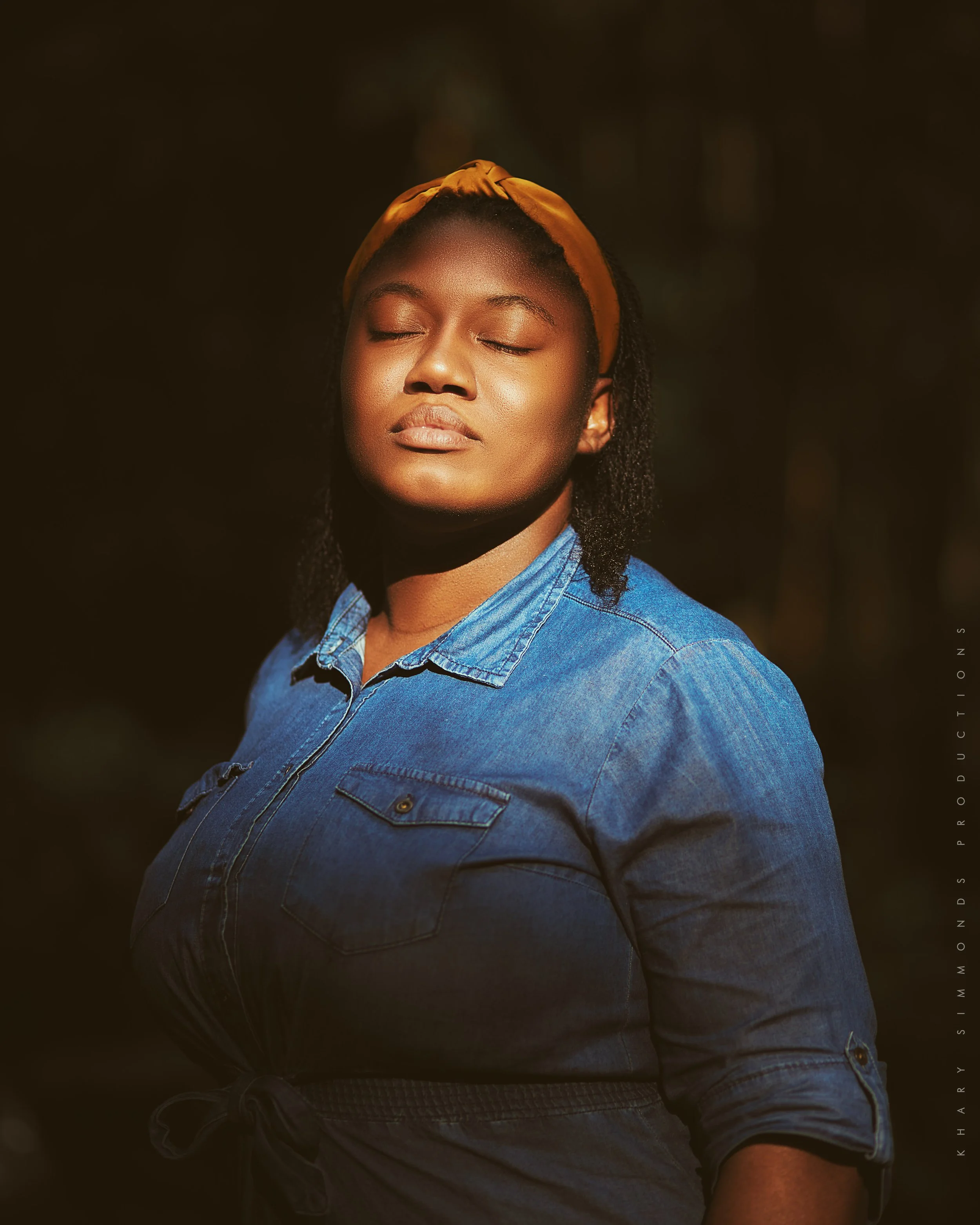 A woman with closed eyes wearing a yellow headband and denim shirt, standing outdoors with sunlight on her face