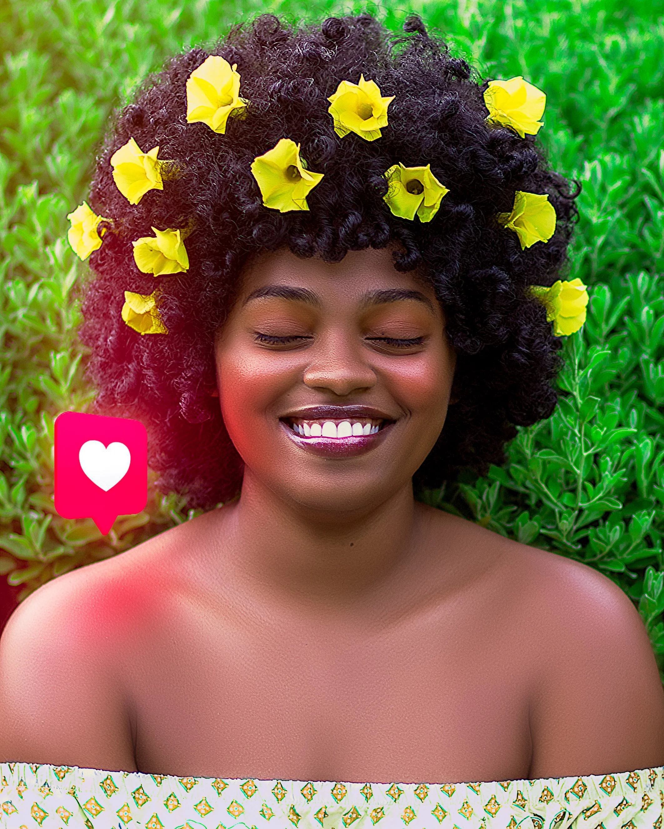 A smiling woman with curly black hair decorated with yellow flowers, standing in front of green foliage.