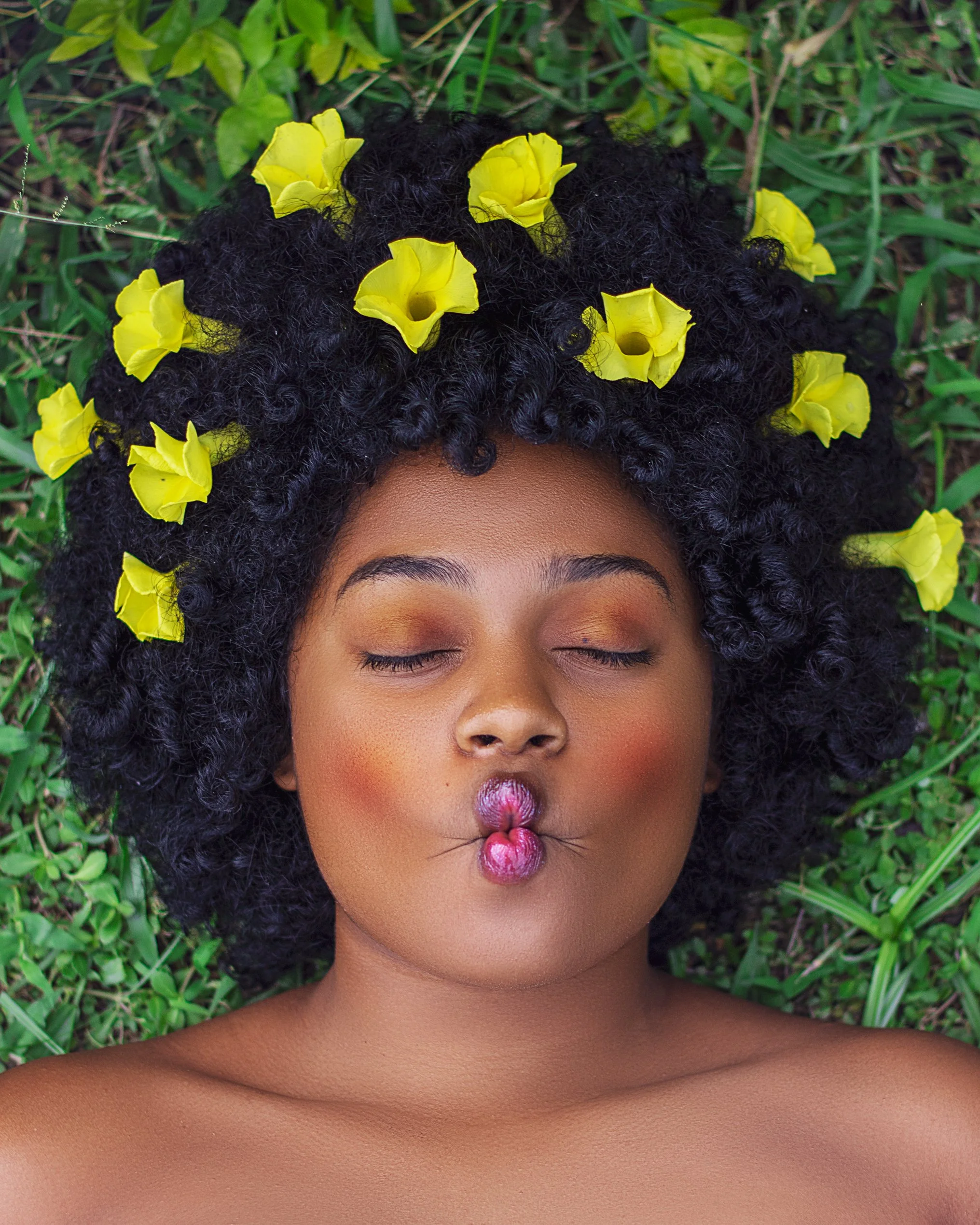 A woman with curly black hair laying on the grass, with yellow flowers arranged in her hair, lips pursed, eyes closed, wearing subtle makeup.