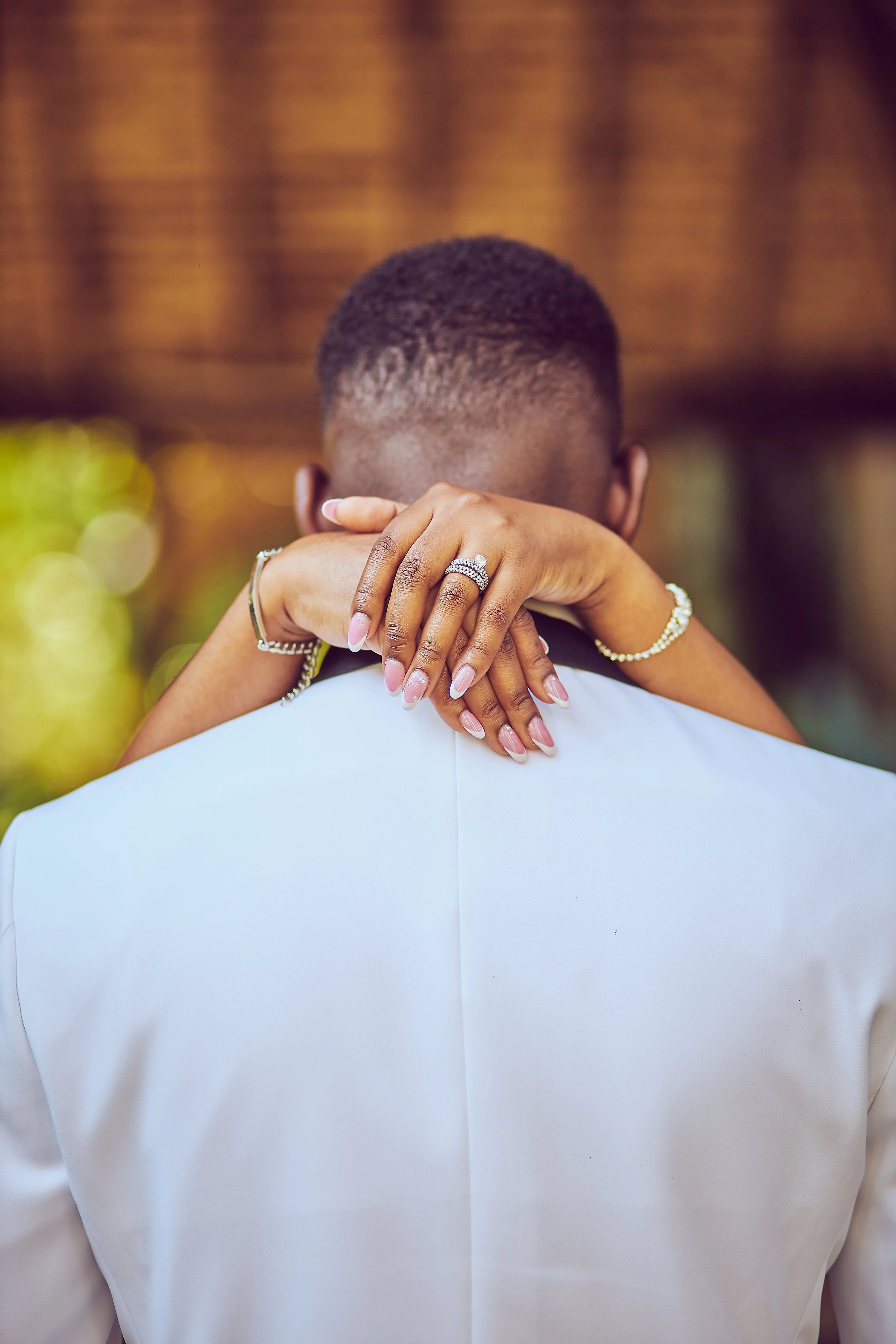 A couple embracing, with the woman’s hands around the man’s neck, showing rings on her fingers. The woman’s nails are manicured, and she is wearing jewelry. The man is dressed in a white shirt, with a blurred background.