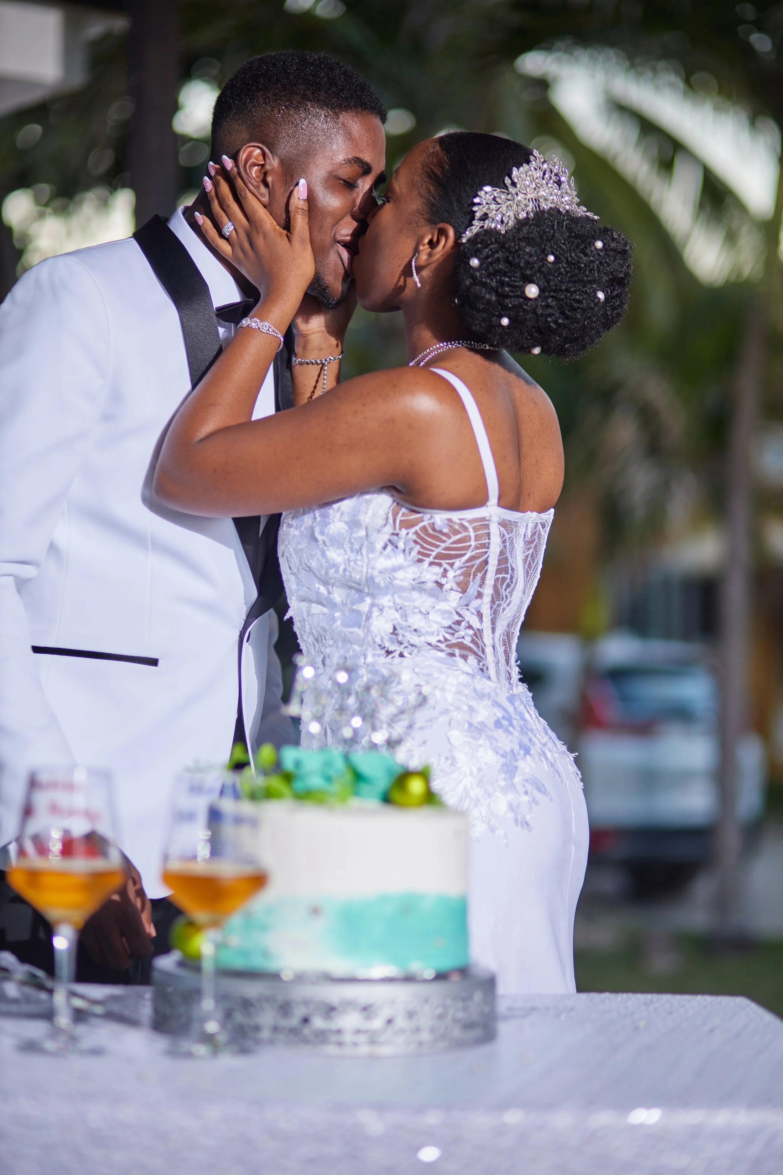 A bride and groom sharing a kiss at their wedding celebration, with a wedding cake and drinks on the table in front of them.