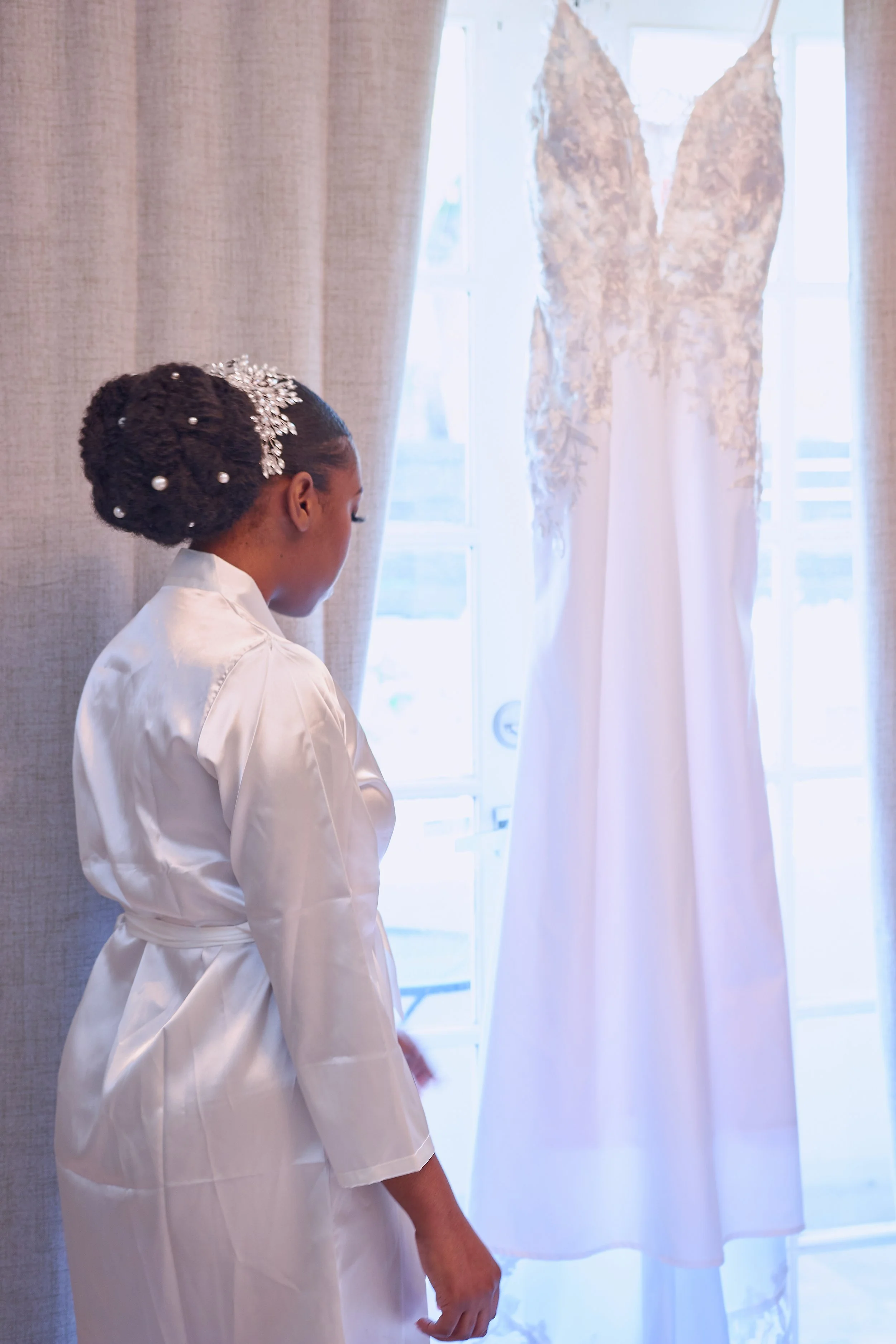 A woman in a satin robe looking at her wedding dress hanging in front of a window.