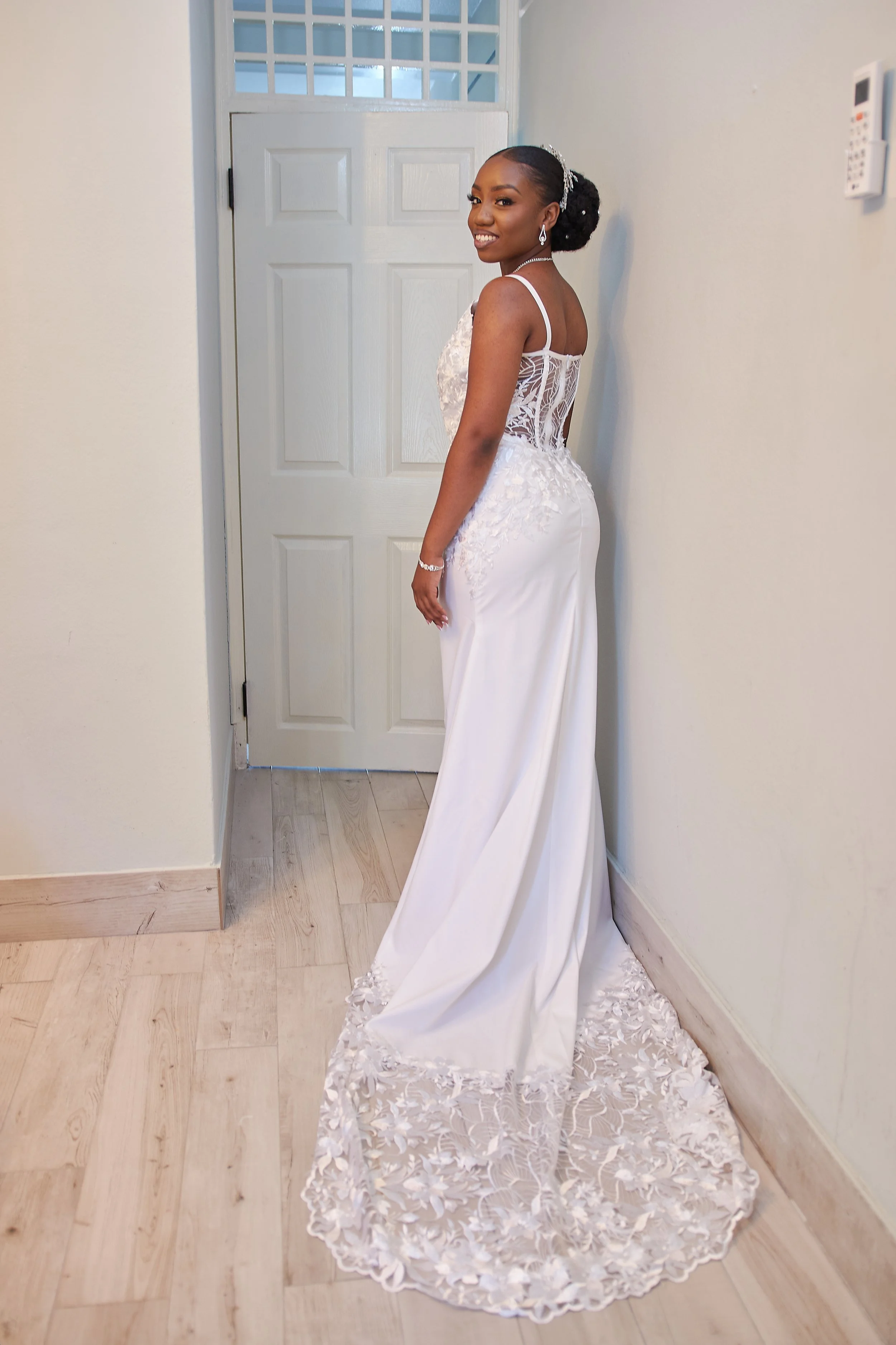 A smiling woman in a white wedding dress with lace details, standing indoors near a closed door and smiling at the camera.