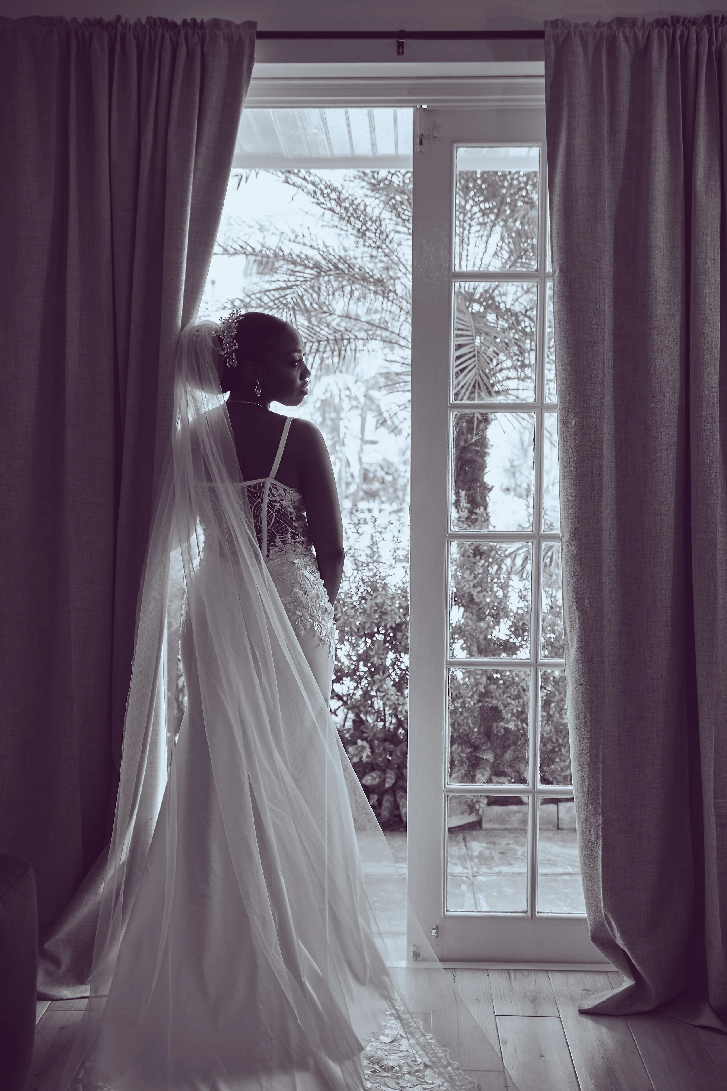 A woman in a wedding dress and veil stands by a glass door, looking outside at trees and plants in a garden.