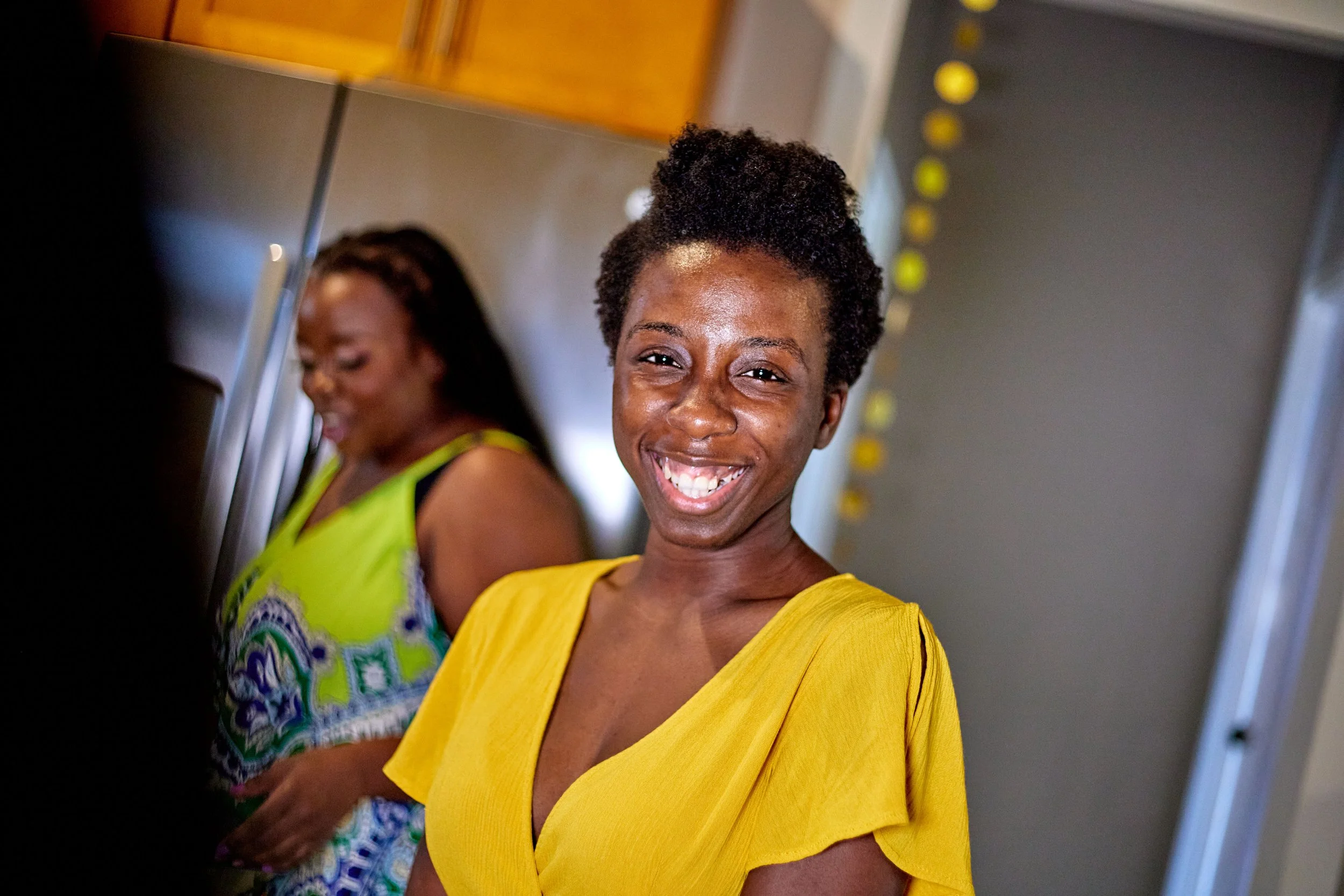 Smiling woman in a yellow shirt, standing indoors with another woman in a colorful dress in the background.
