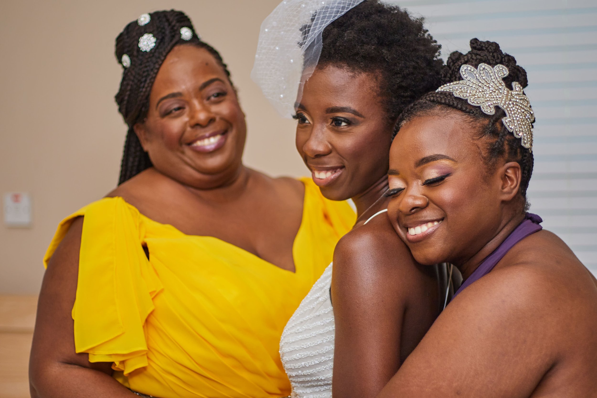 Three women, two in colorful dresses and one in a white bridal gown, happy and embracing at a celebration or wedding, indoors.