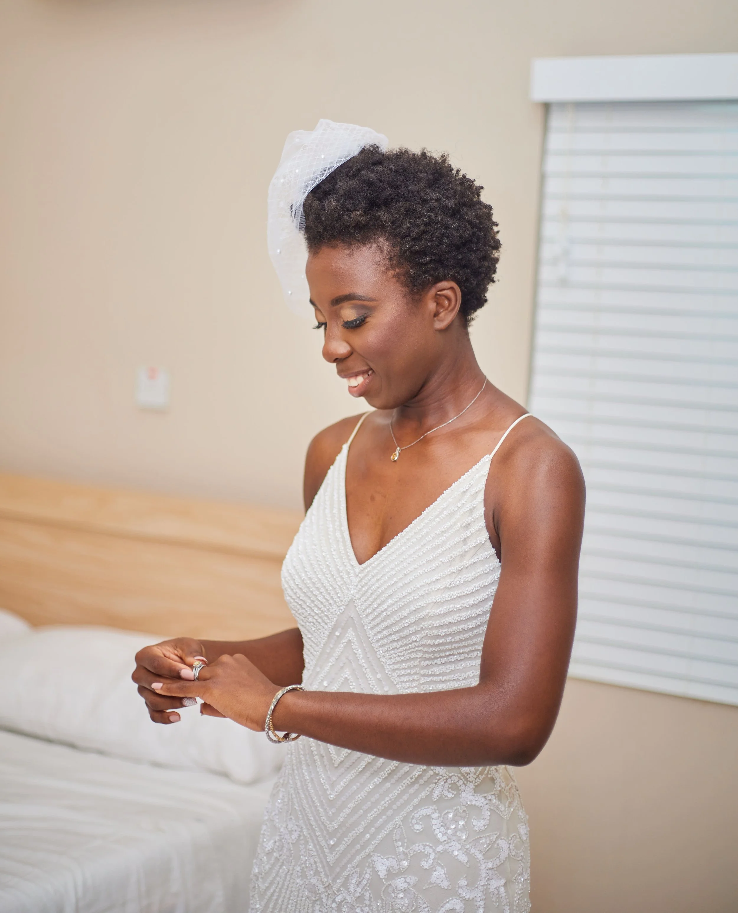 A woman in a white wedding dress with beaded details standing in a room with a beige wall, white window blinds, and a bed, smiling and looking down.