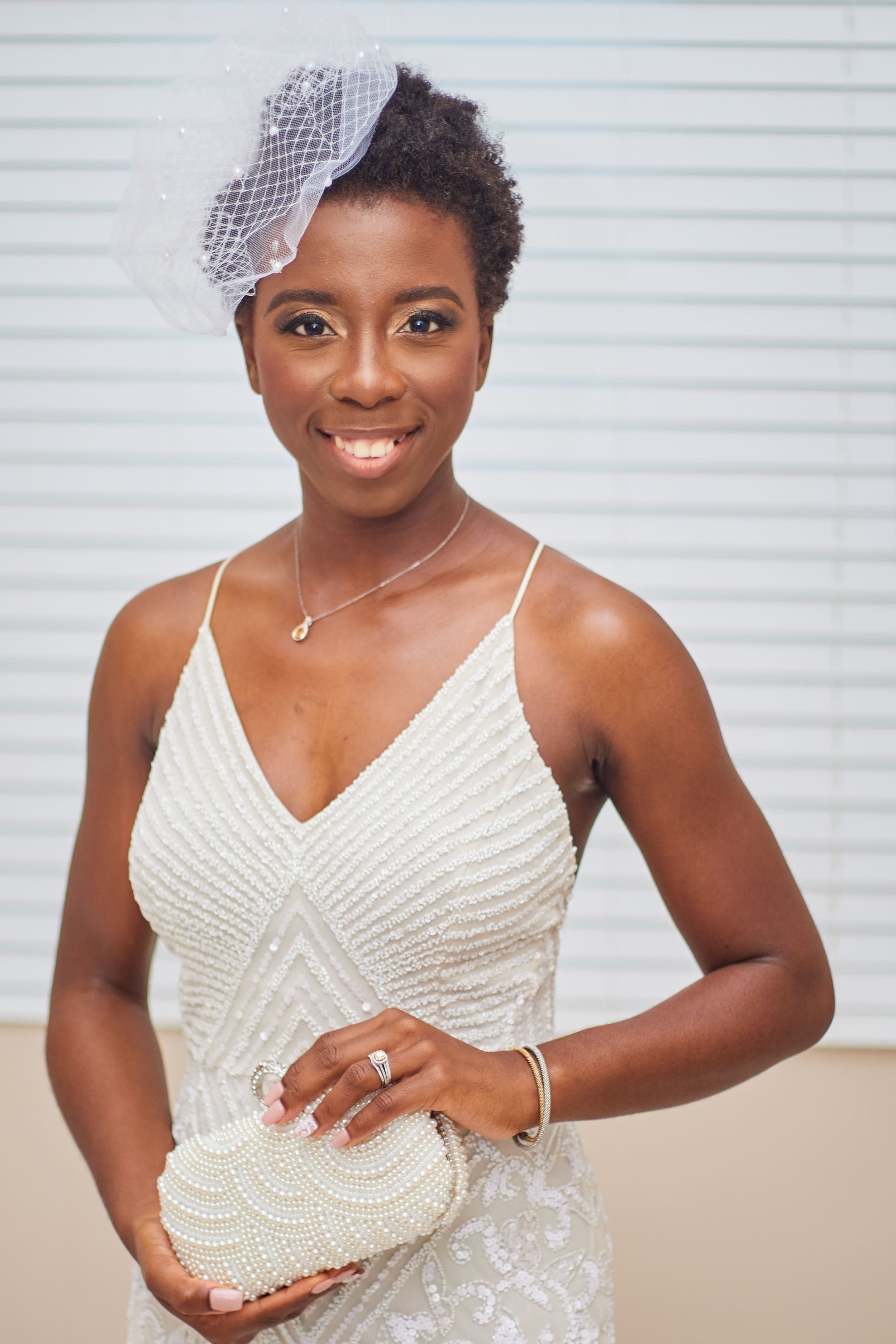 A woman in a white dress, wearing a headpiece and jewelry, holding a pearl embellished clutch, smiling in front of window blinds.