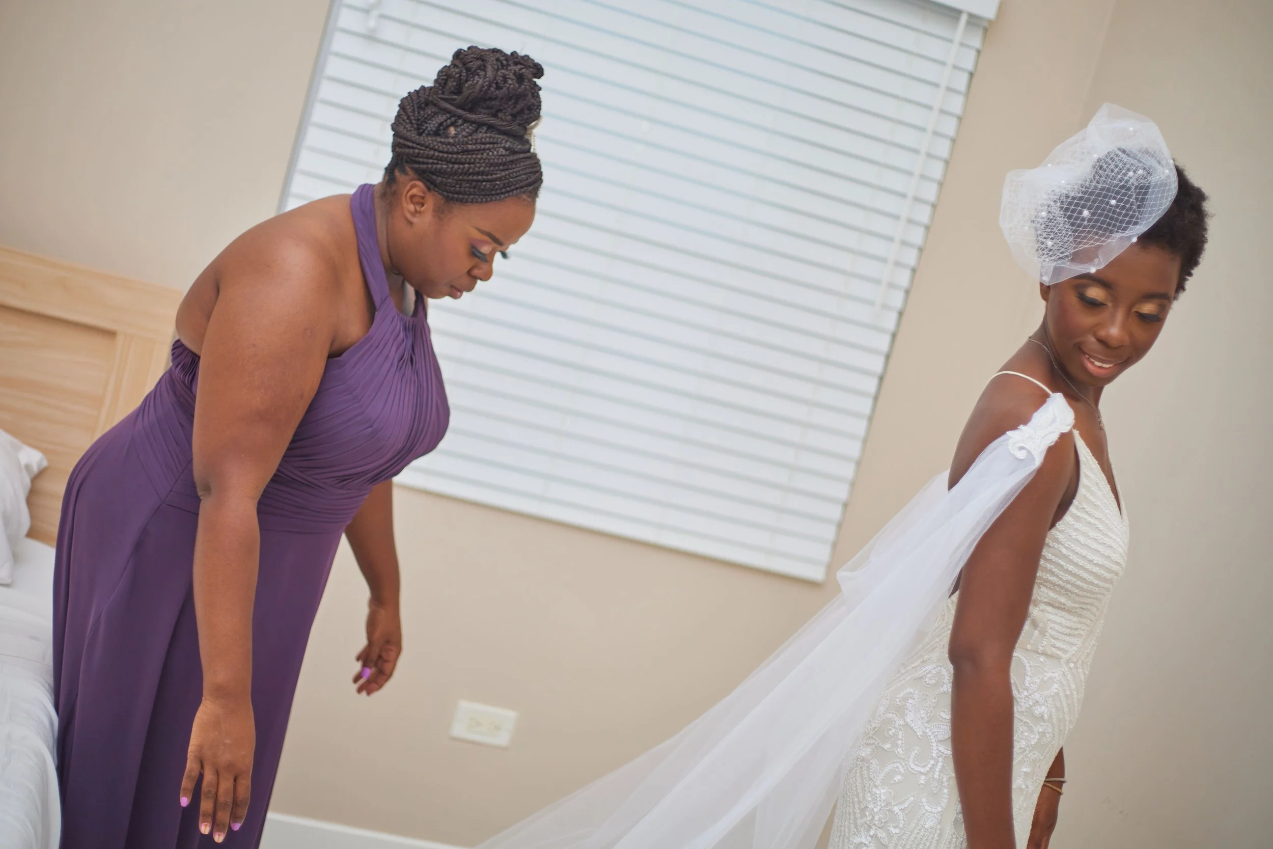 A bride in a white wedding dress with a veil, smiling, and another woman in a purple dress preparing in a room with beige walls and a window with white blinds.
