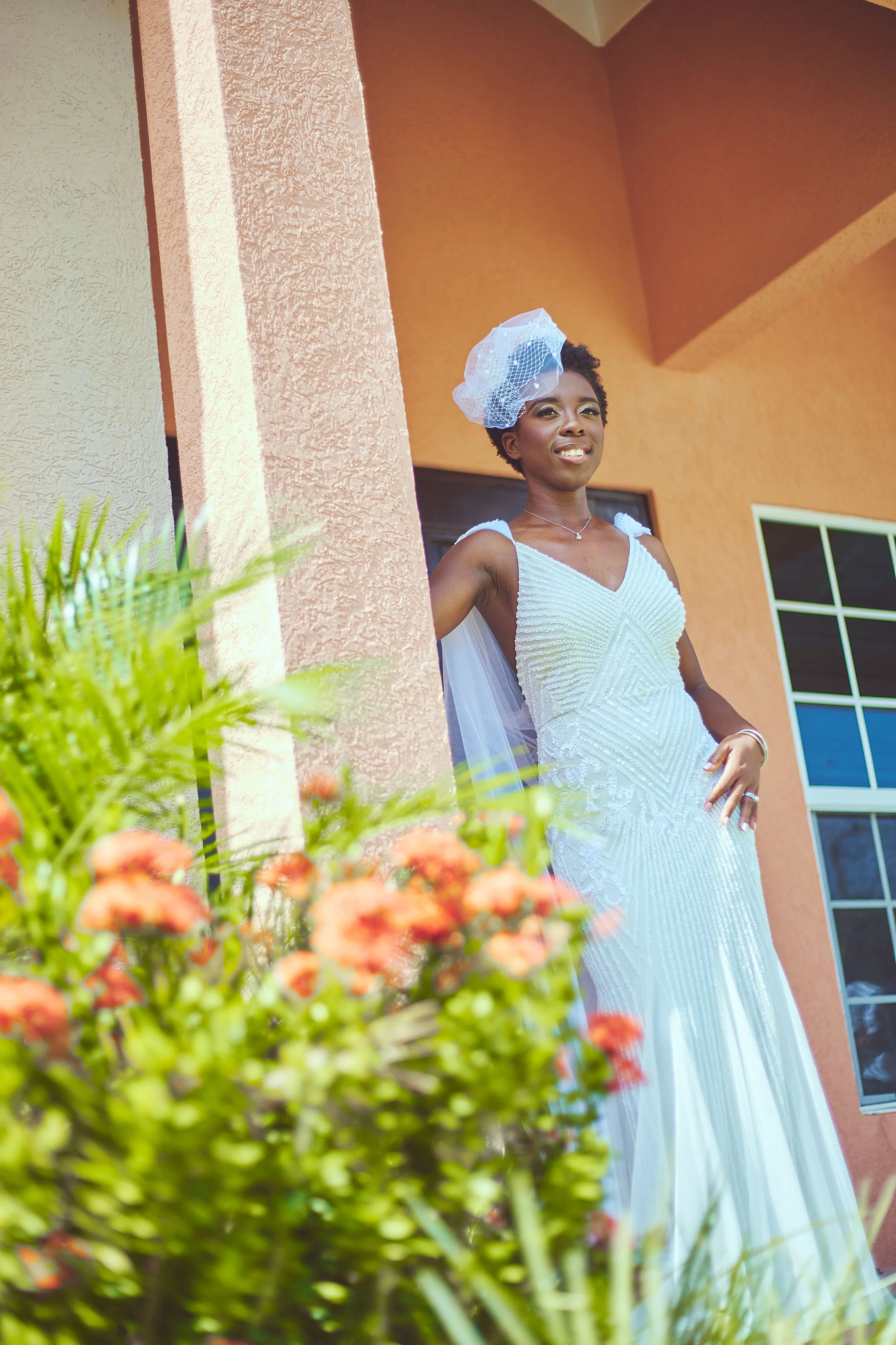 A woman standing outdoors in a white dress with a tulle headpiece, smiling, with flowers and a house in the background.