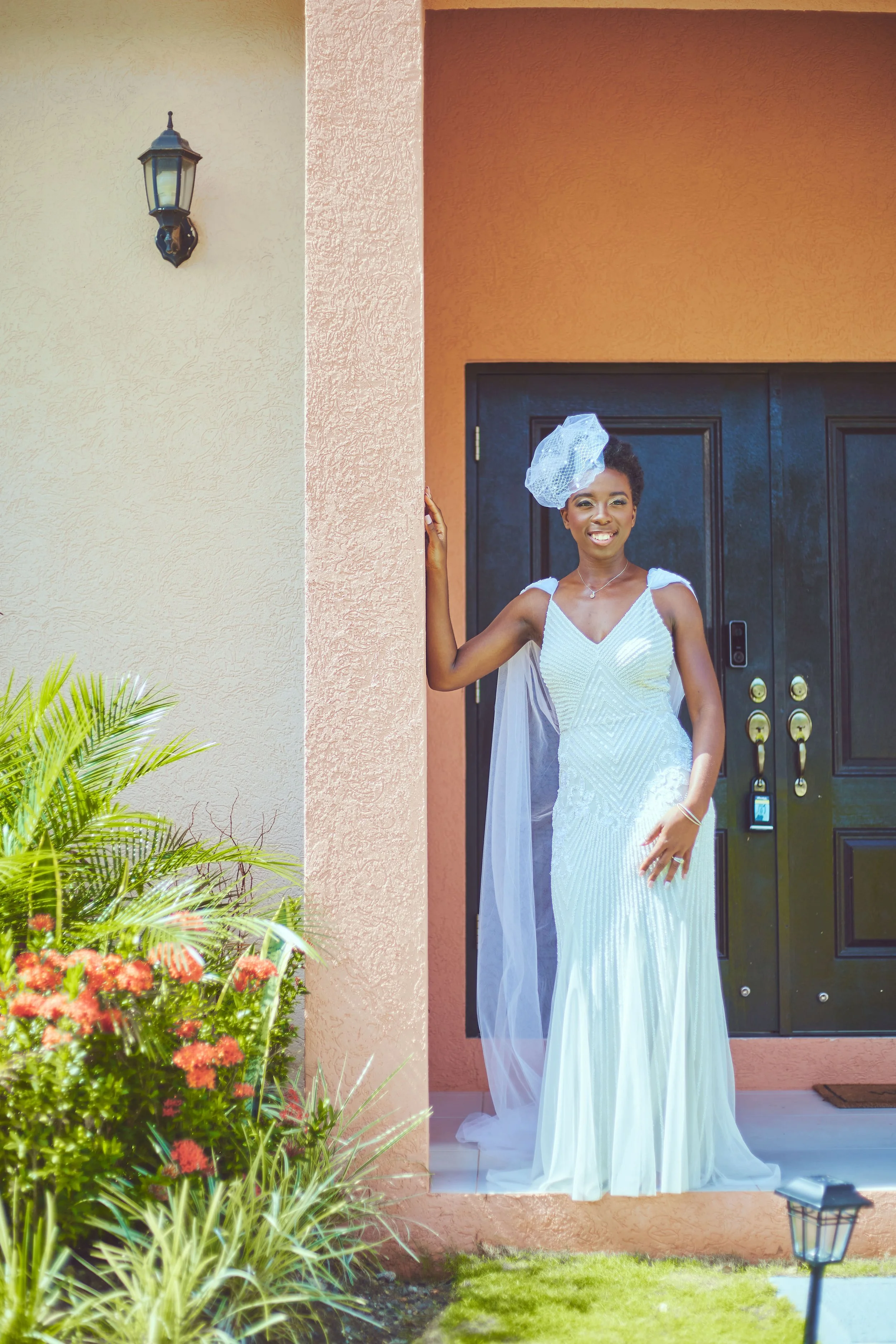 A woman in a white dress and a fascinator hat standing at the entrance of her house, smiling, with plants and flowers to the side.